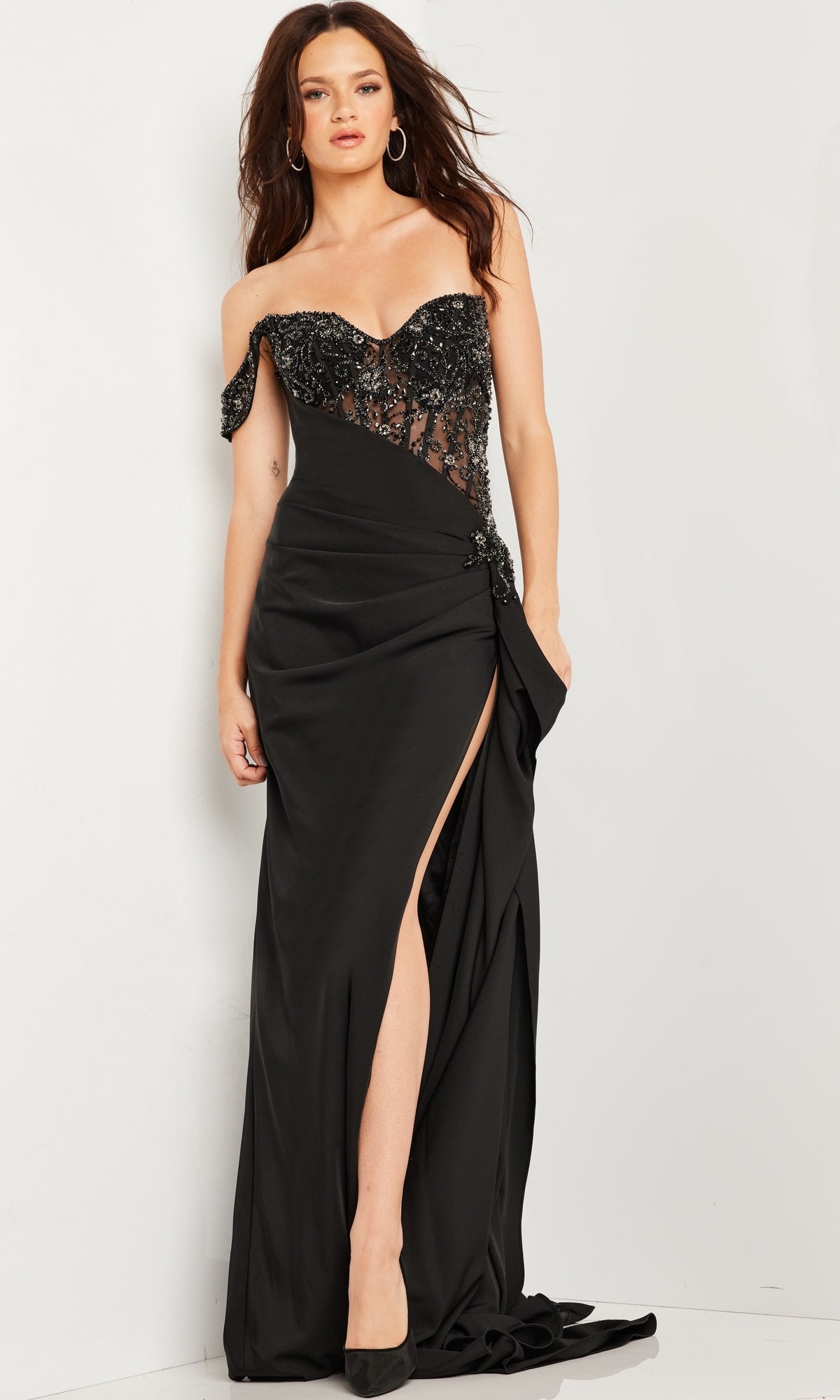 A woman stands against a white background wearing the Long Prom Dress 37094 by Jovani—an off-the-shoulder black gown with a sheer beaded bodice, high slit, and black heels.