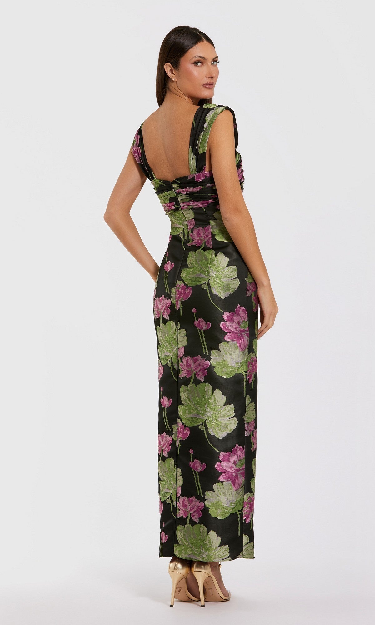 Wearing the Mac Duggal 37095, a fitted, floor-length black brocade formal dress with green and purple floral print and an open back, paired with gold high heels, this woman stands against a white background.
