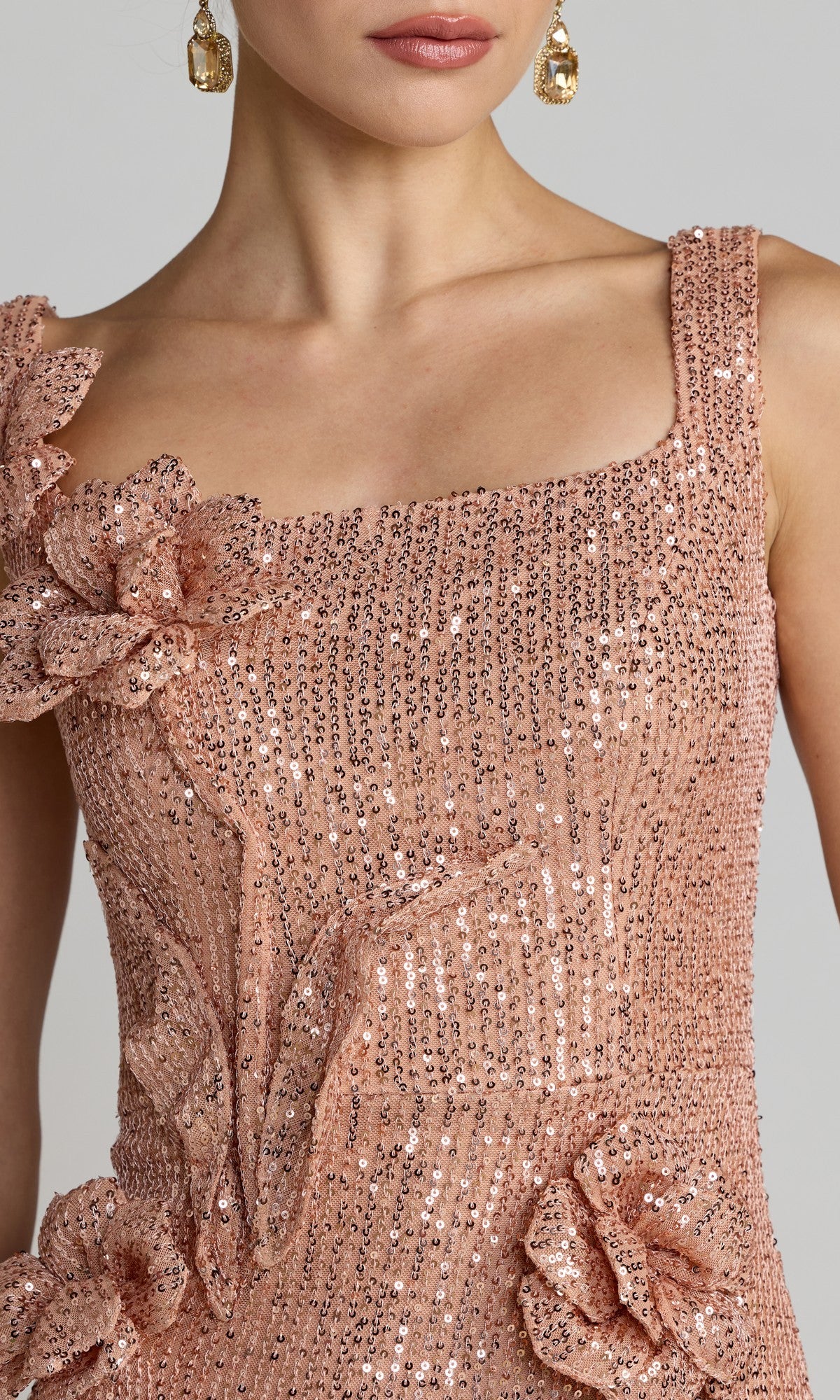 Wearing the Mac Duggal 37104 long formal dress, a woman stuns in blush pink with a sleeveless, square neckline silhouette. The dress features shimmering sequins and bold fabric flower accents. She completes her look with gold drop earrings and subtle, neutral makeup.