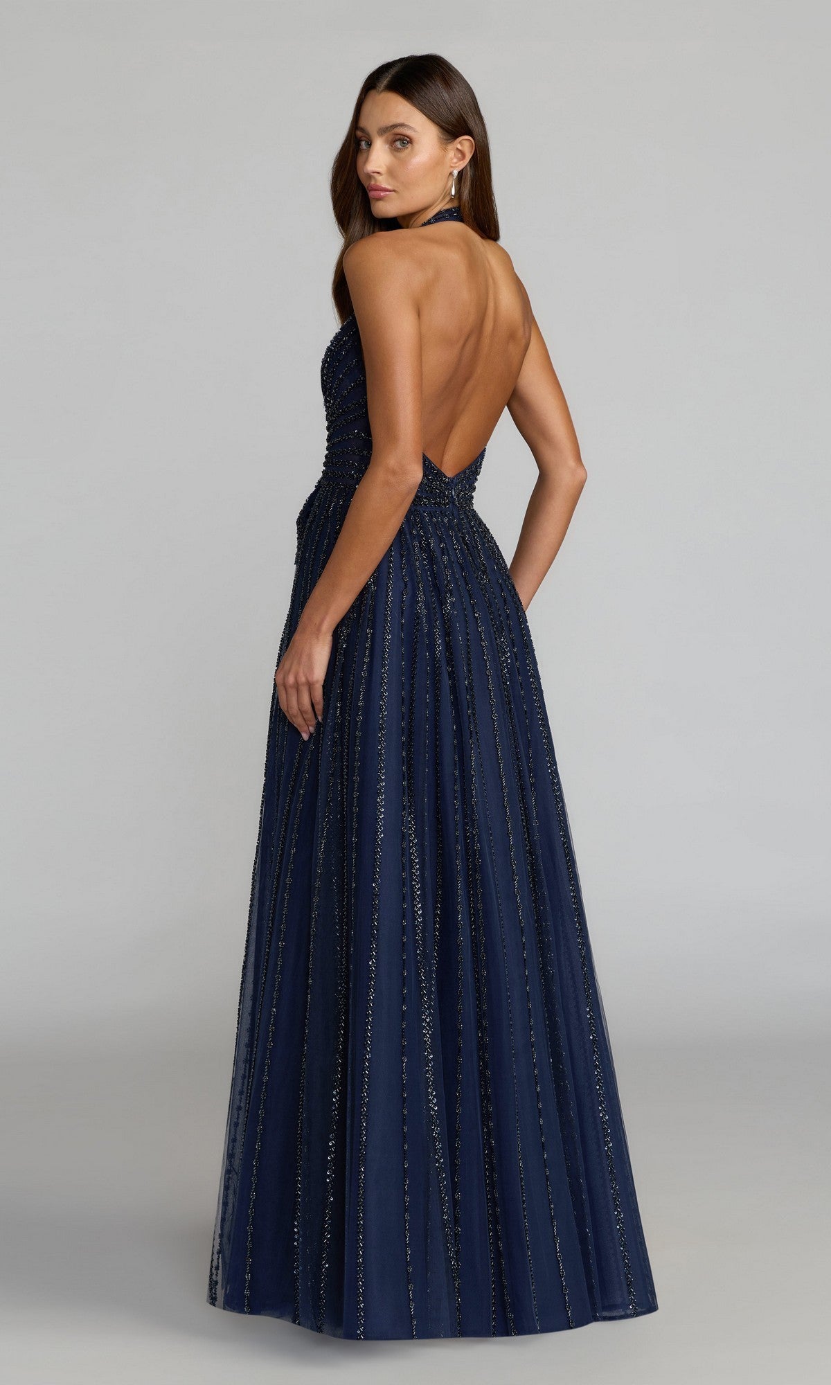 A woman poses with her back to the camera in the Mac Duggal 37120 long formal dress, a floor-length navy halter gown featuring sparkly vertical stripes.
