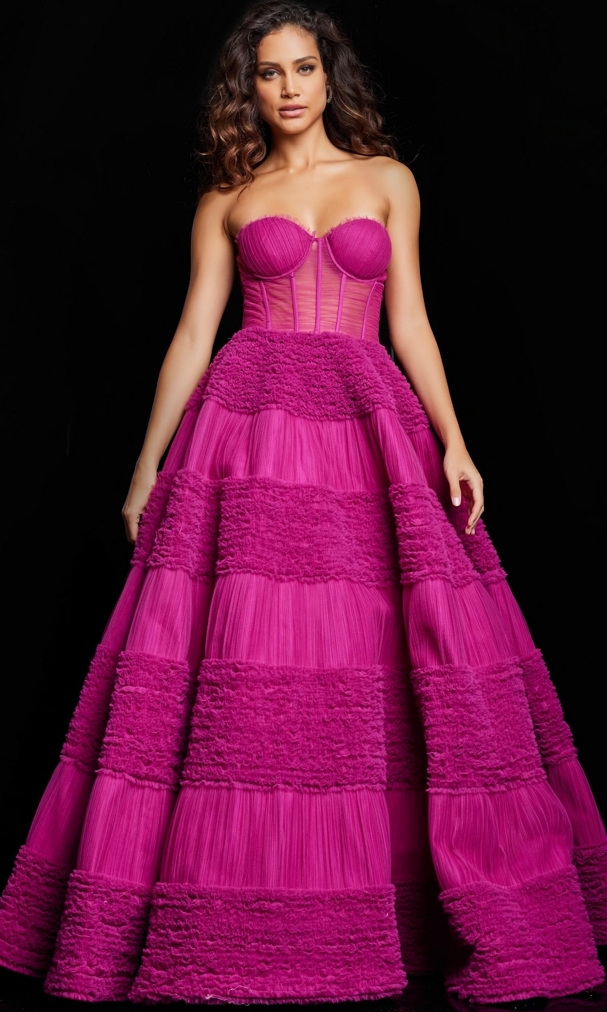 A woman poses elegantly against a black background in the Strapless Sheer-Corset Jovani Ball Gown 37157, featuring a sweetheart A-line bodice and a tiered, textured magenta skirt.