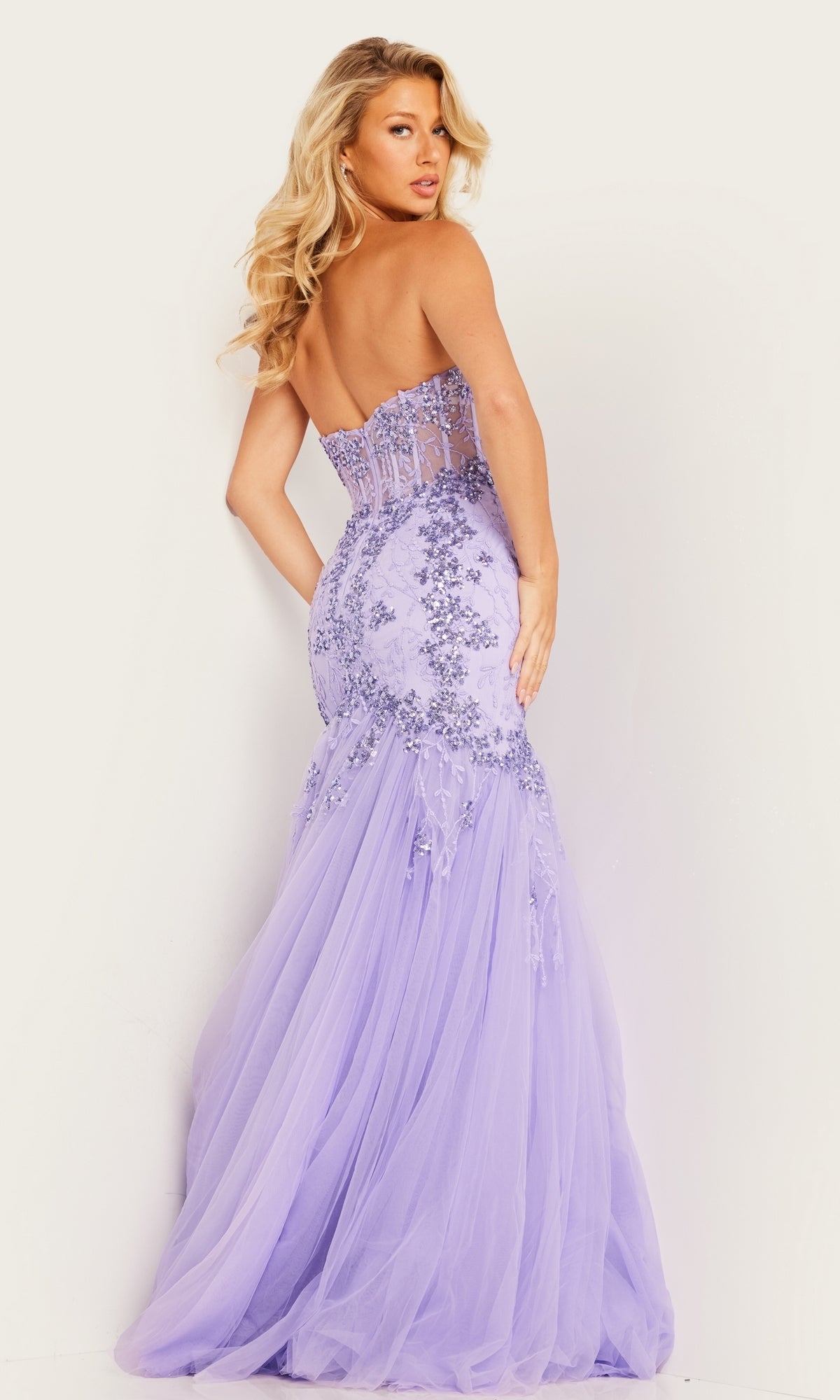 A woman poses with her back to the camera in the Jovani Long Prom Dress 37249, a strapless sweetheart lavender gown featuring lace detailing and a tulle mermaid skirt—a stunning choice for prom.