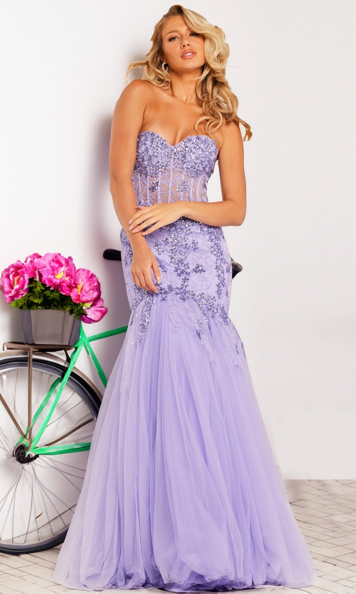 A woman in the Long Prom Dress 37249 by Jovani, featuring a strapless sweetheart mermaid design with floral embroidery, stands beside a bicycle decorated with pink flowers.