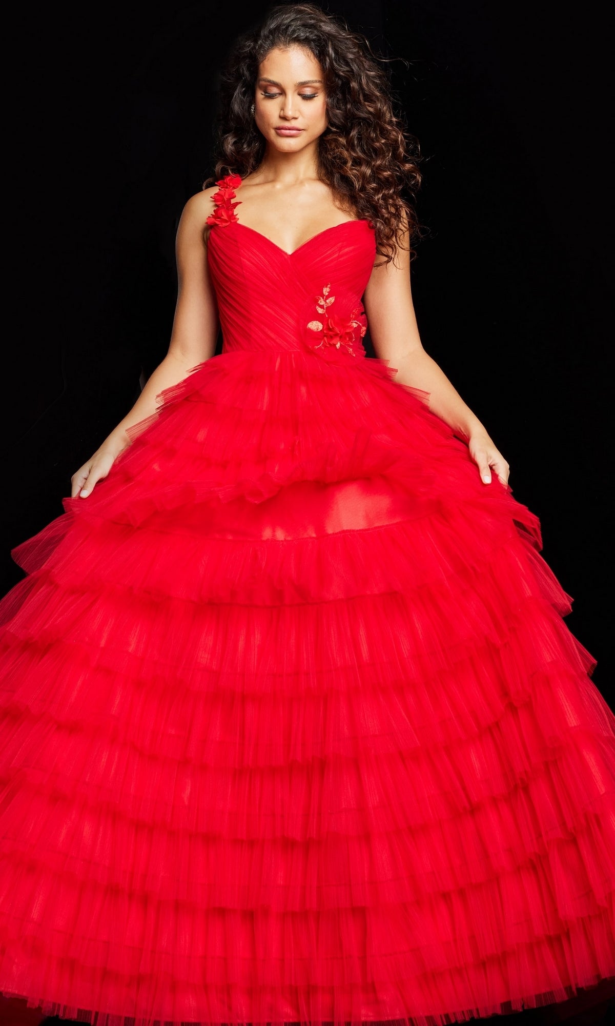 A woman in the Jovani Long Prom Dress 37274—a vibrant red ruffle ball gown with layered tulle and floral embroidery on the bodice—stands against a black background.