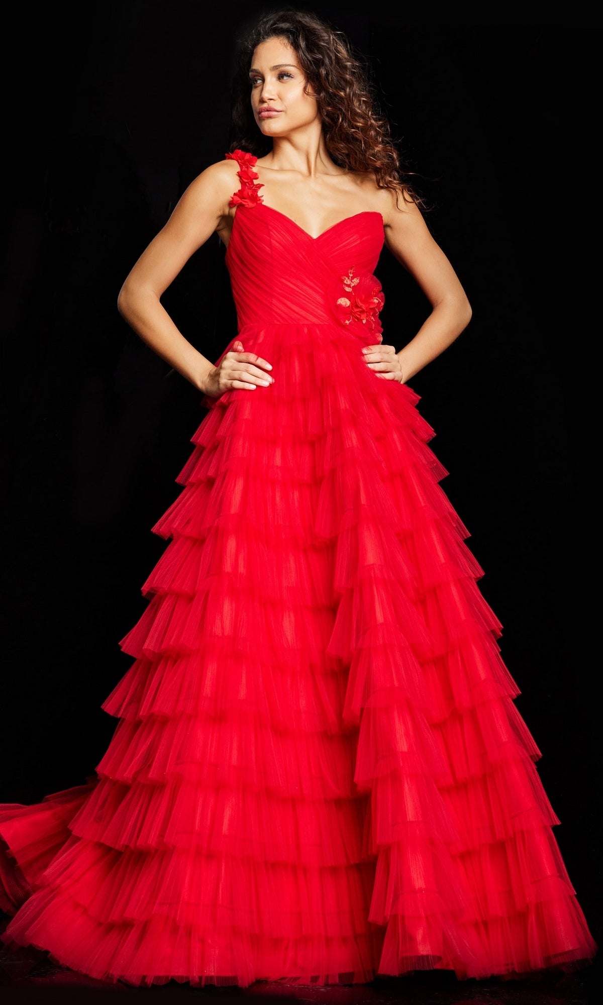 A woman poses against a black background, wearing the Long Prom Dress 37274 by Jovani—a floor-length, one-shoulder red ball gown with multiple tulle ruffles and floral appliqué on the bodice.