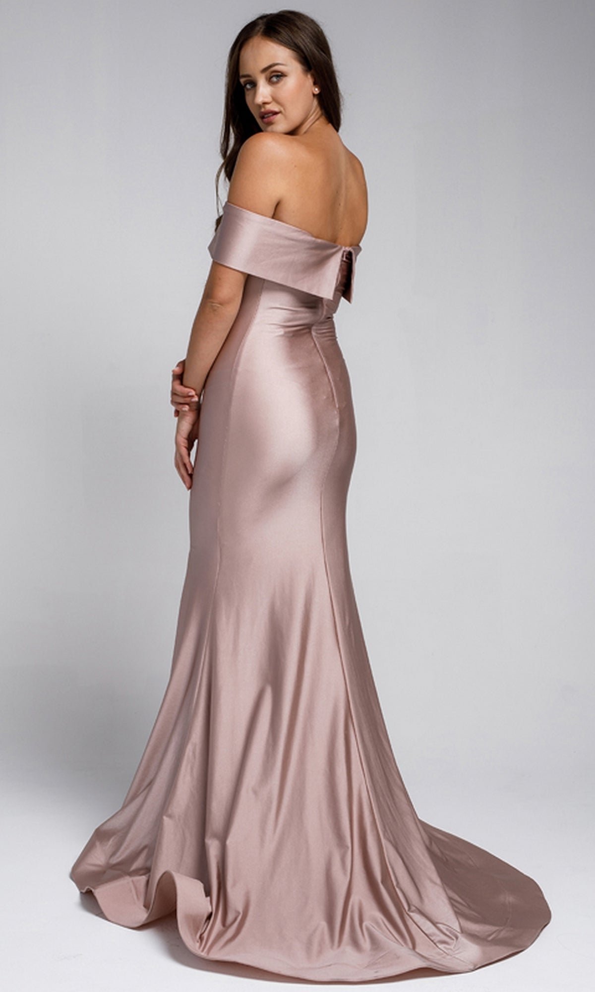 Woman in the Sleek Simple Off-the-Shoulder Long Prom Dress 373, a floor-length, light pink satin gown, stands and looks over her shoulder against a plain background.