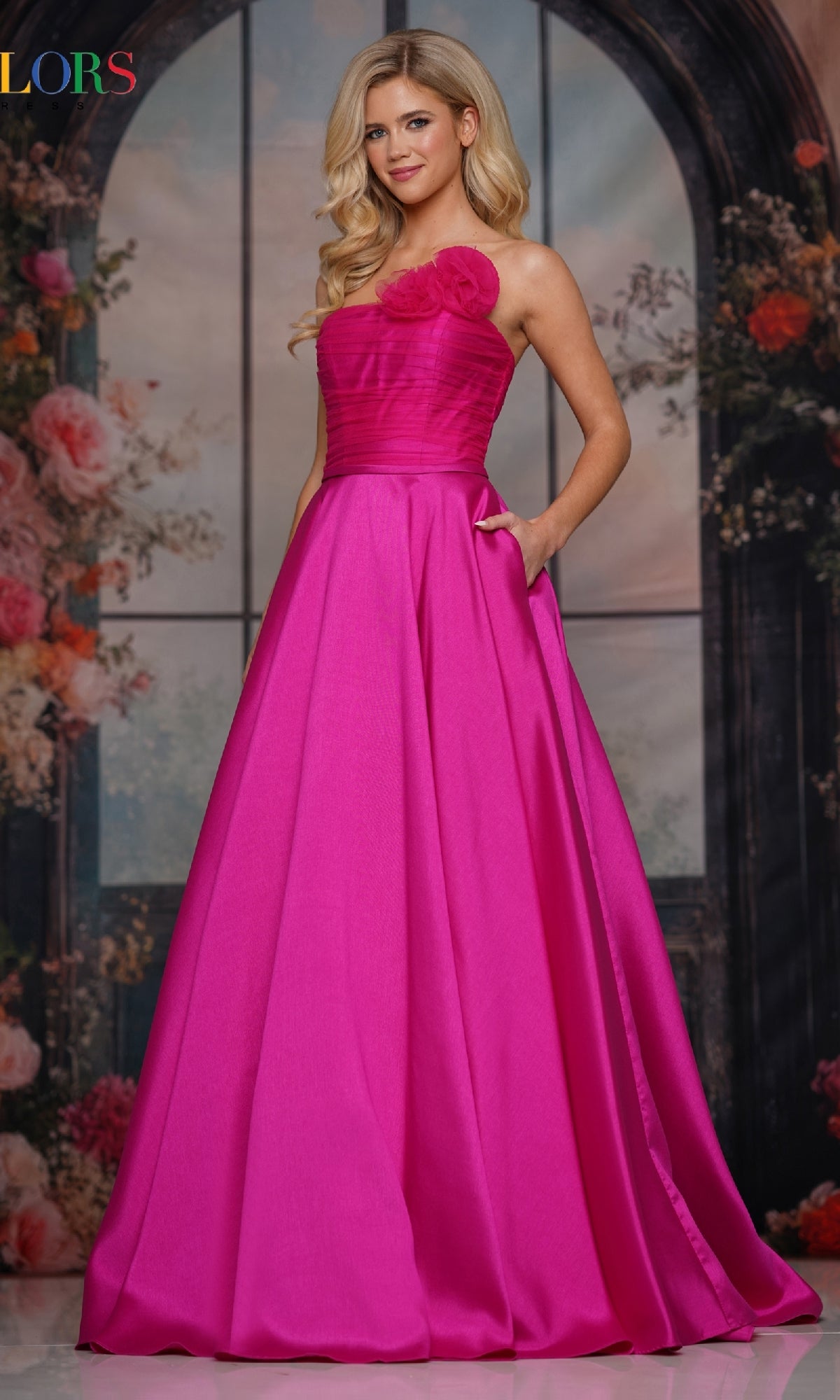 A woman stands before a floral backdrop in the magenta Strapless Long A-Line Prom Dress: Colors Dress 3737, featuring a strapless neckline, floral bodice detail, and pockets for an elegant prom or evening look.