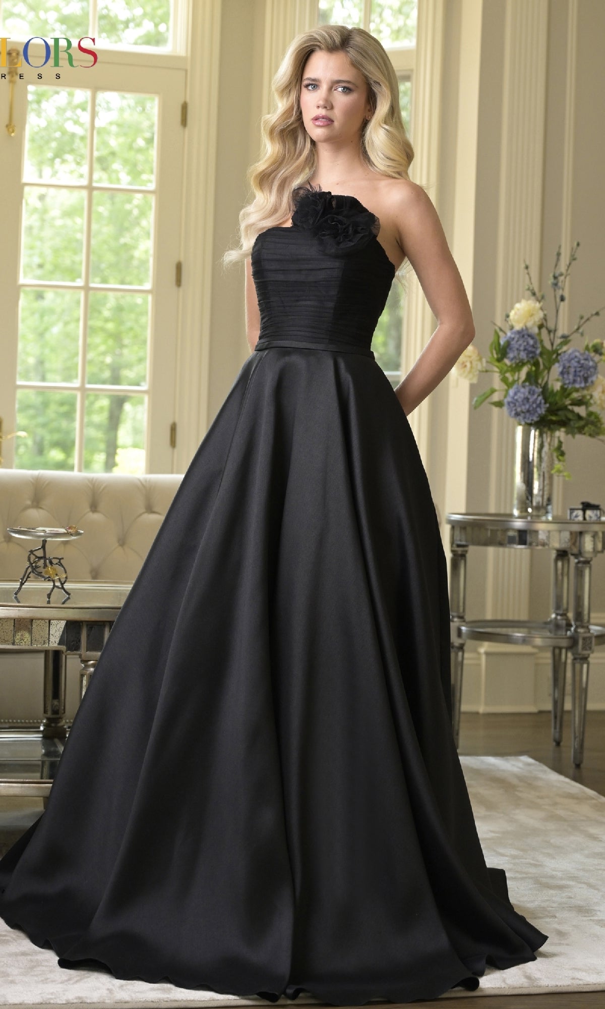 A woman wearing the Colors Dress 3737, a strapless long A-line prom dress, stands in an elegant living room filled with light, featuring large windows and floral arrangements.