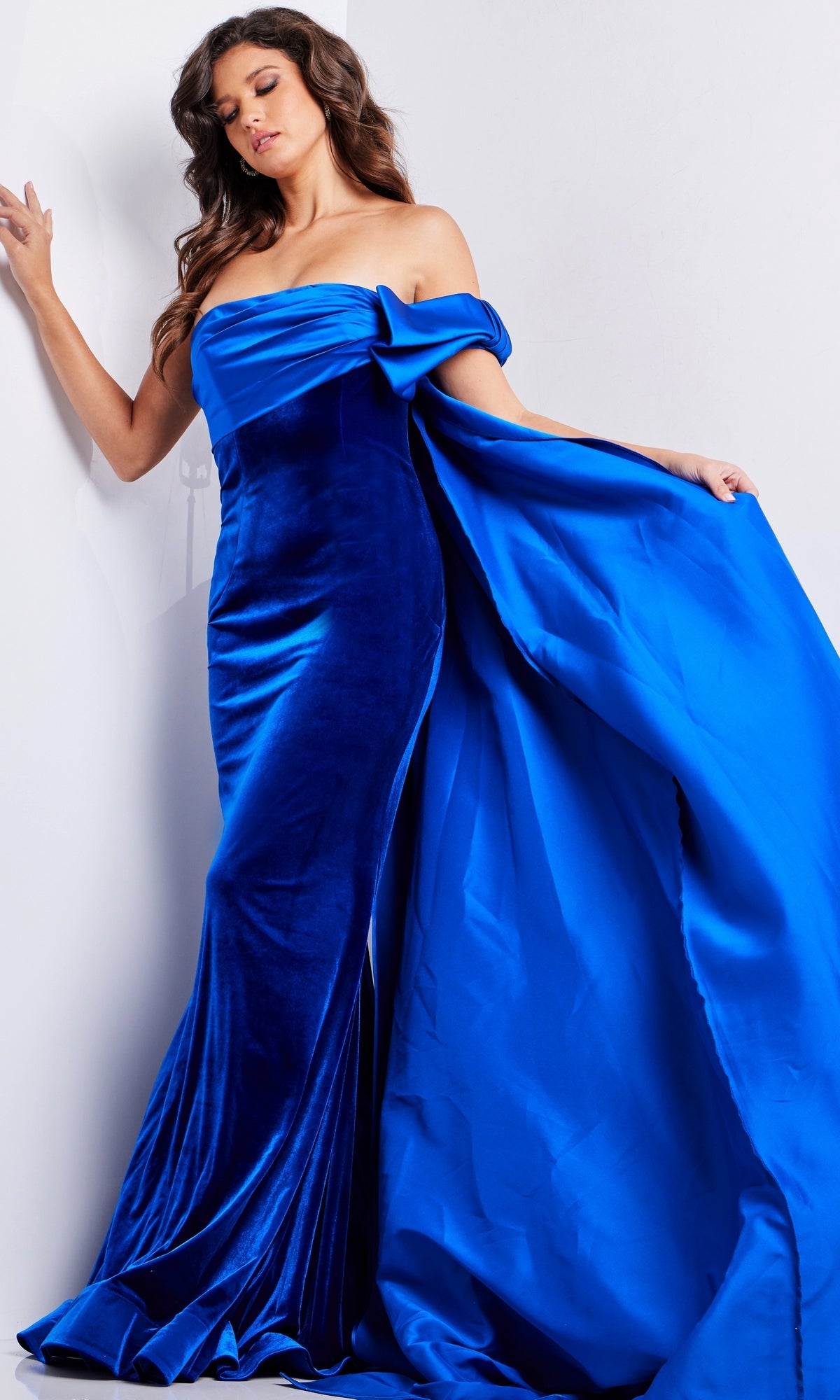 Woman in the Long Prom Dress 37390 by Jovani, a royal blue velvet fitted gown with an off-the-shoulder neckline and dramatic train, posing against a plain white background.