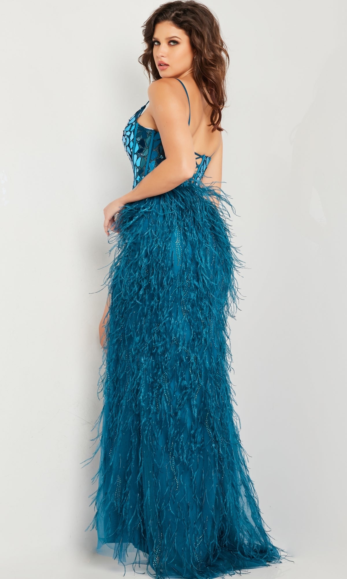 A woman models the Jovani 37435 Feather Long Sequin Prom Dress, featuring a fitted teal silhouette with spaghetti straps, lace-up back, and feathered skirt detailing, against a plain light background.