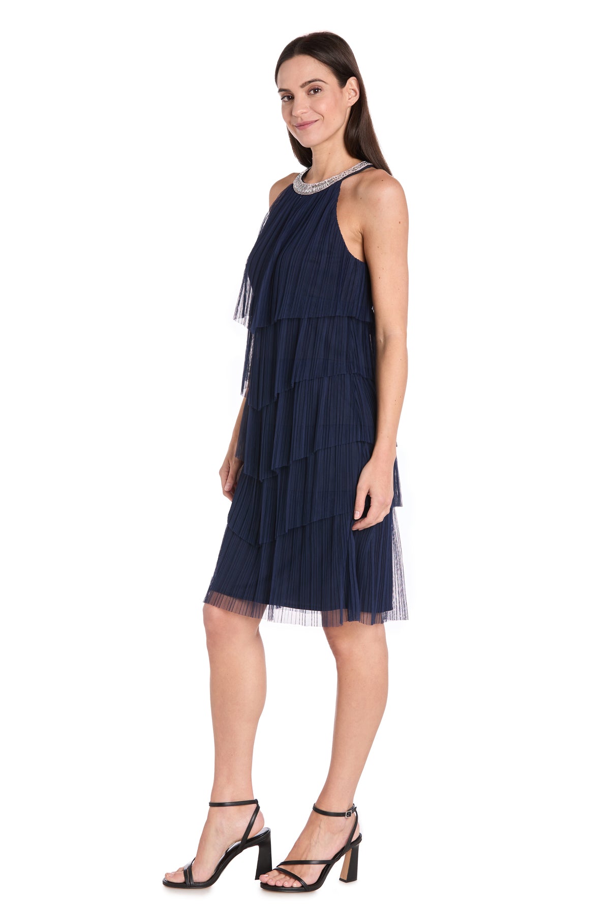 A woman in profile wears the R&M Richards 3744 Short Navy Blue Cocktail Dress with pleated tiers and chiffon ruffles, paired with black heeled sandals, standing against a white background.