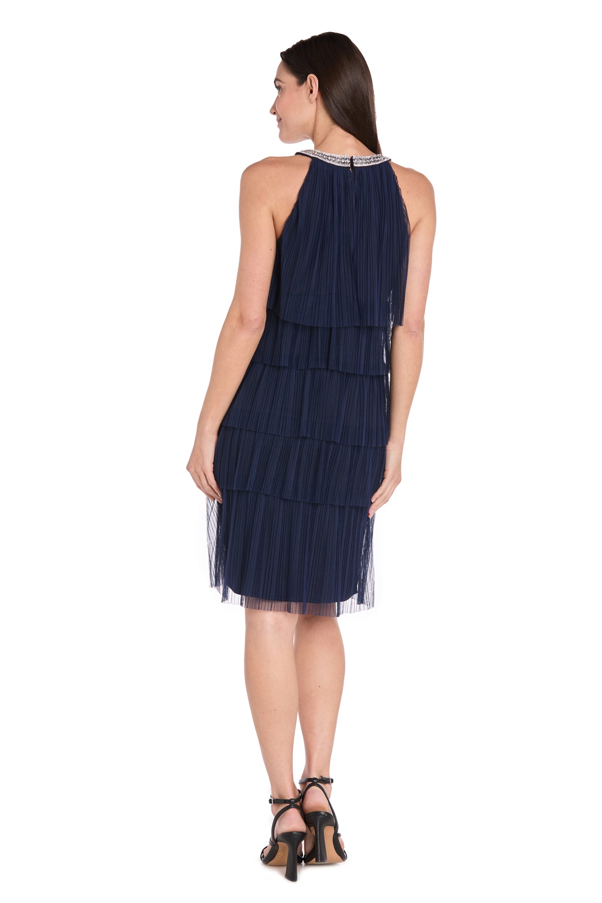A woman stands facing away, wearing the R&M Richards 3744P Petite Short Navy Party Dress with a layered knee-length design and black high-heeled sandals, set against a plain white background.