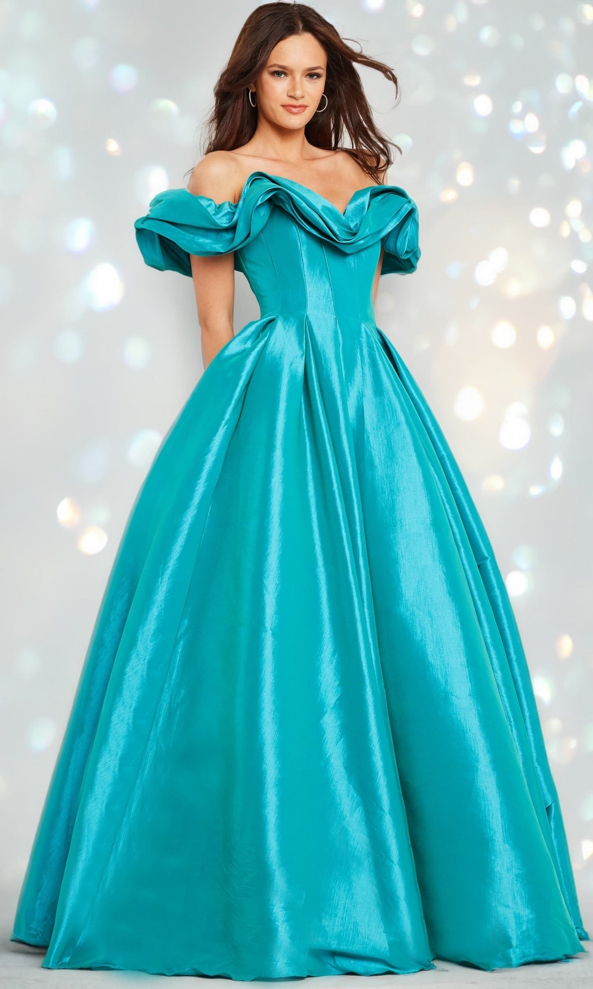 A woman stands wearing the Long Prom Dress 37476 by Jovani, featuring a teal hue, off-the-shoulder ruffled neckline, and a full skirt, set against a softly lit background.