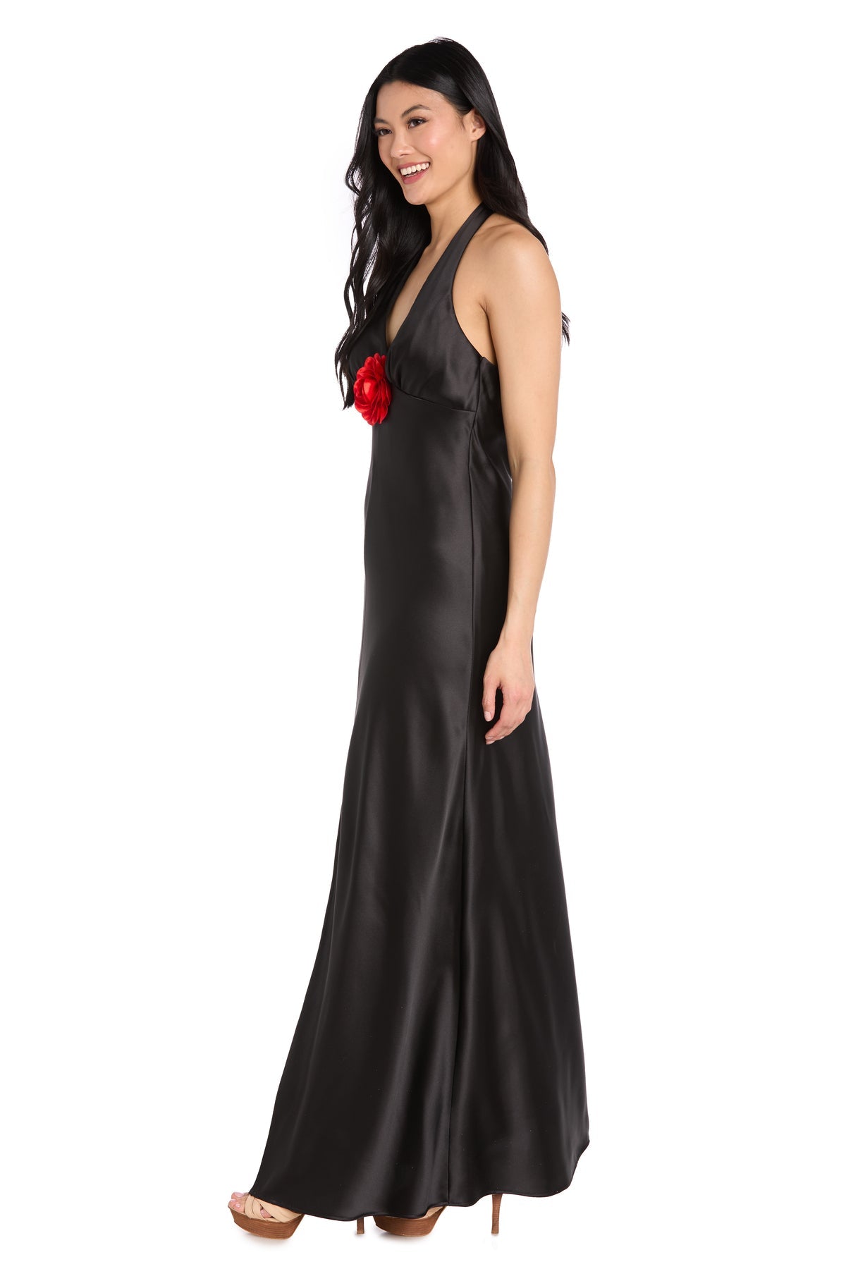 Woman in the R&M Richards 3773 Black Satin Halter Formal Dress with red flower chest detail stands in high heels, smiling against a white background—ideal for any black-tie event.