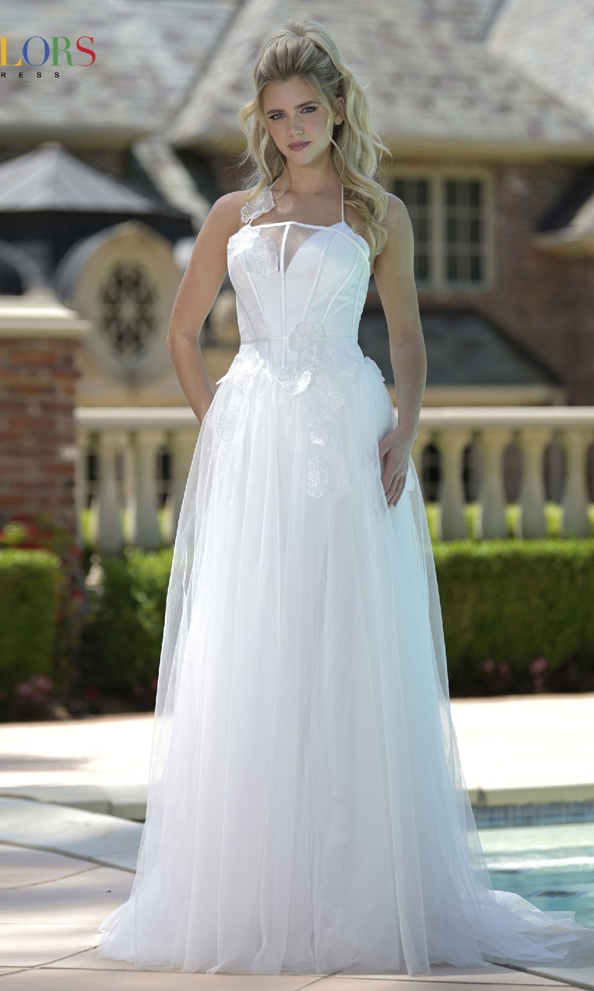 A woman in the Colors Dress 3796, a white halter-neck long prom dress with 3D floral appliques, stands outdoors in front of a building and greenery.