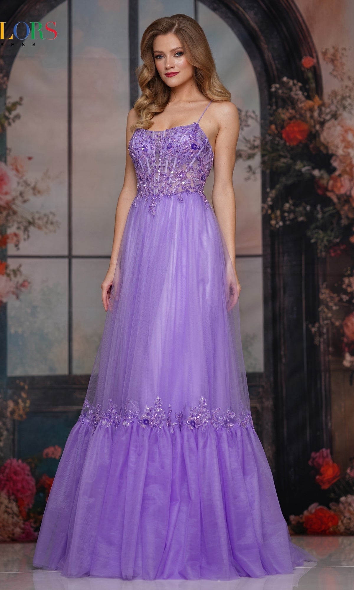 A woman in the Colors Dress 3799, a floor-length prom dress with a sleeveless embroidered sheer bodice and floral accents, poses before a floral backdrop with arched windows.