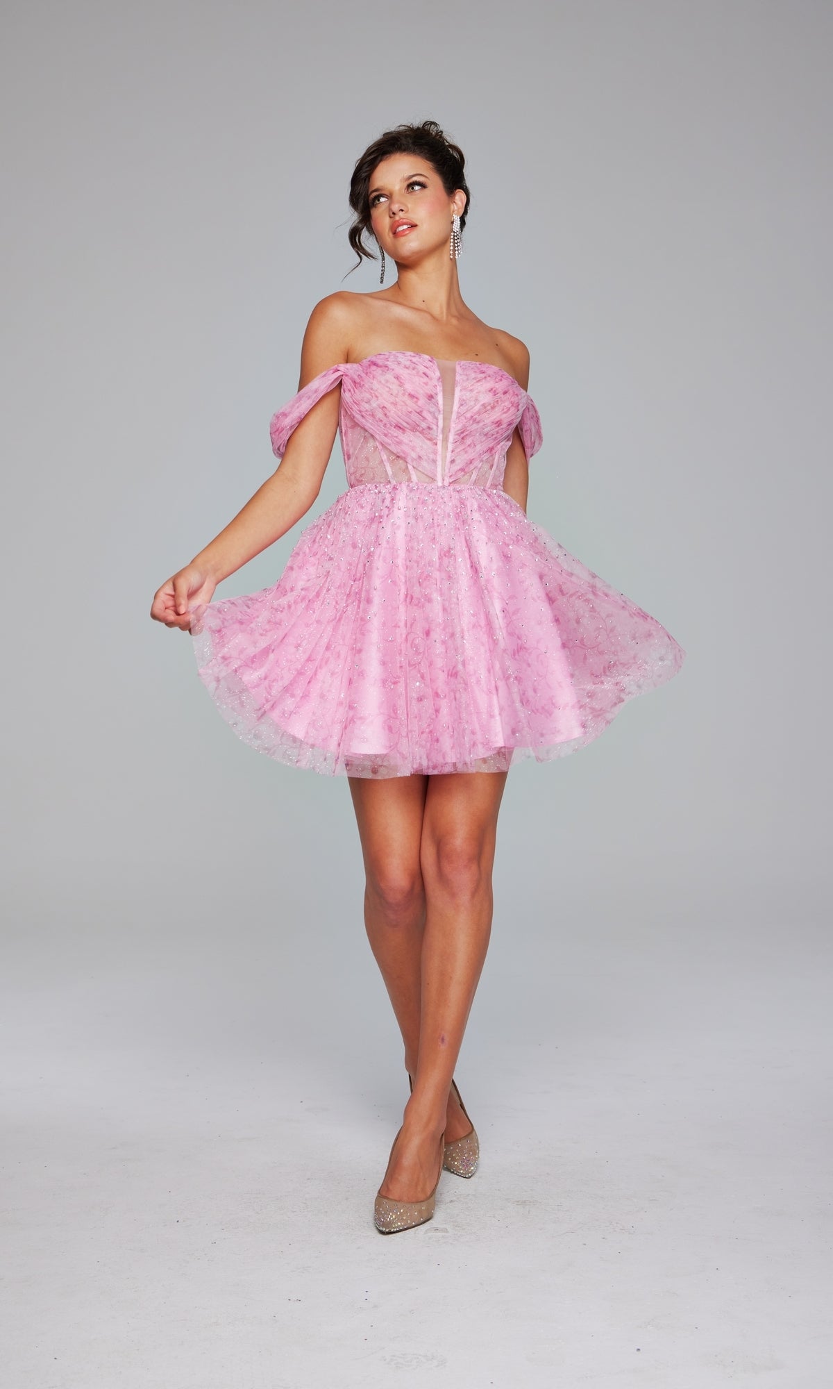 A woman poses against a plain light gray background wearing the Jovani Off-Shoulder Short Pink Hoco Dress 38000 with floral patterns, sheer panels, and sparkly heels.