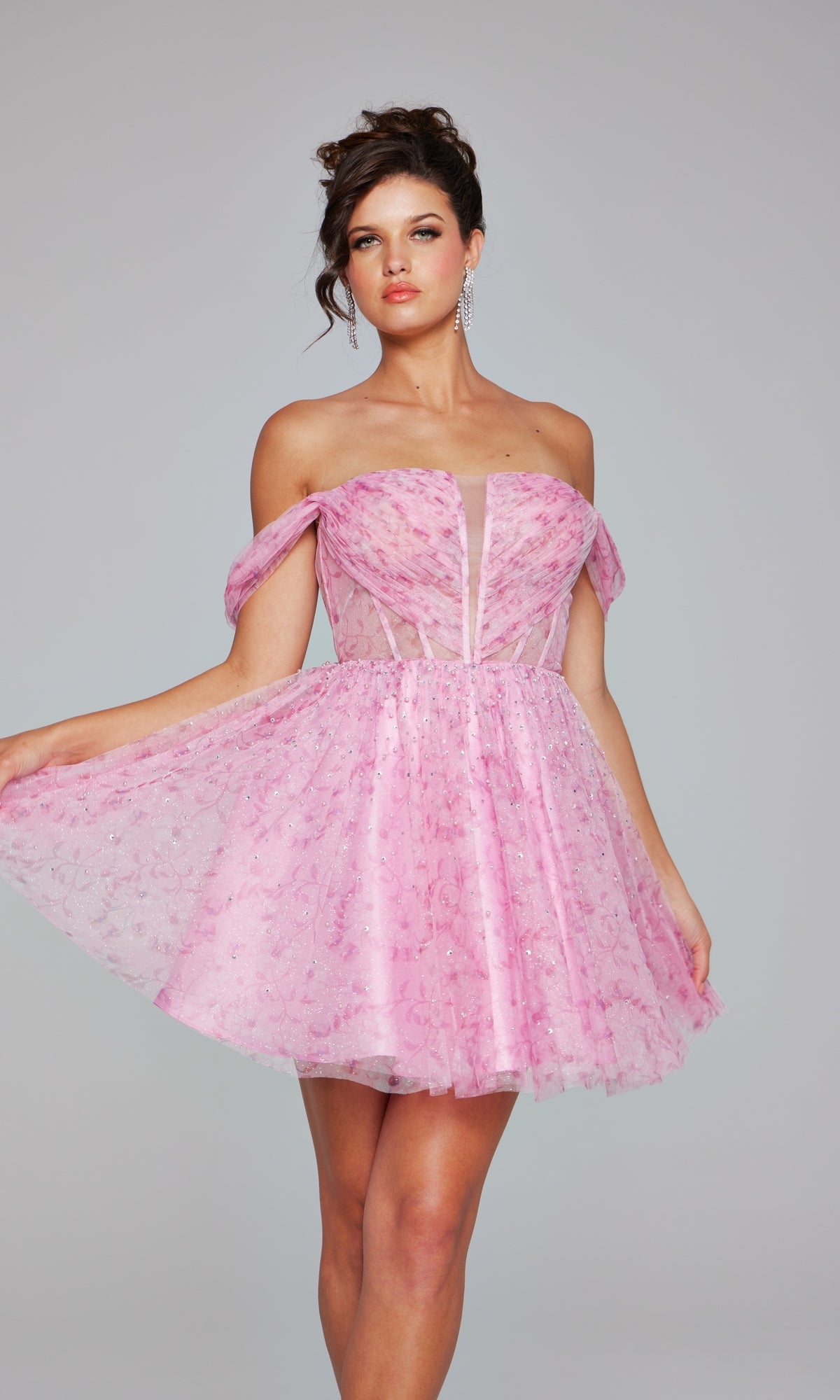 A woman models the Jovani Off-Shoulder Short Pink Hoco Dress 38000—a chic pink lace designer dress with sheer accents—against a plain gray background, showcasing elegance and style.