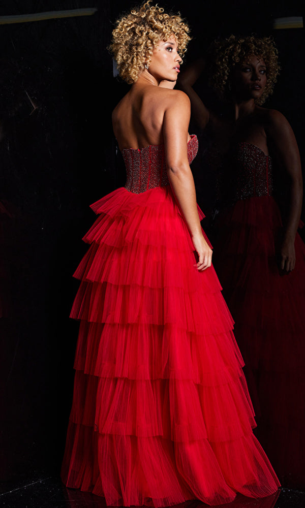A woman with curly hair wears the Jovani Long Prom Dress 38090, a strapless ball gown with layered red tulle, standing before a mirror against a dark background.