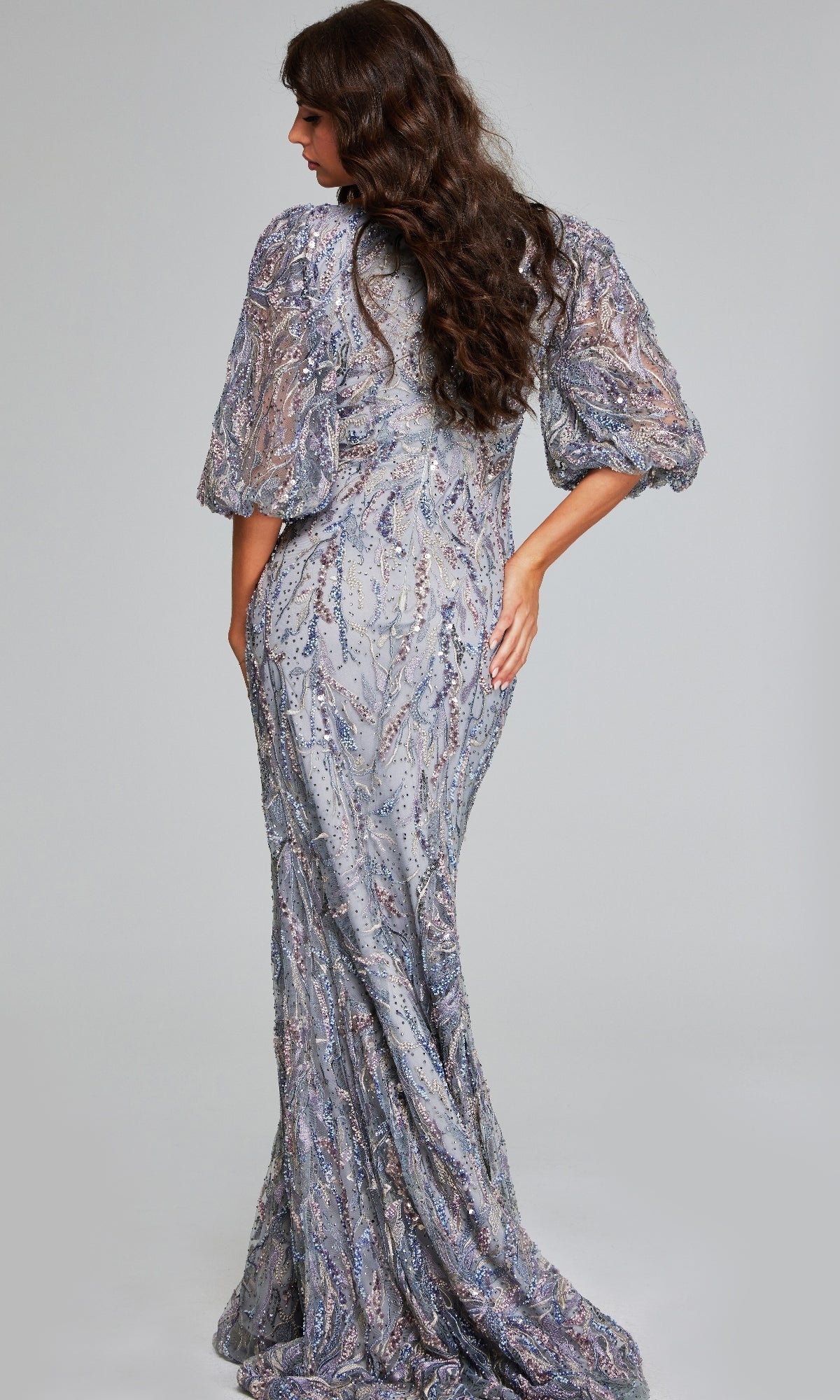 A woman with long wavy hair faces away, hand on hip, wearing the Jovani 38110 Long Grey Formal Dress adorned with sparkly embellishments.