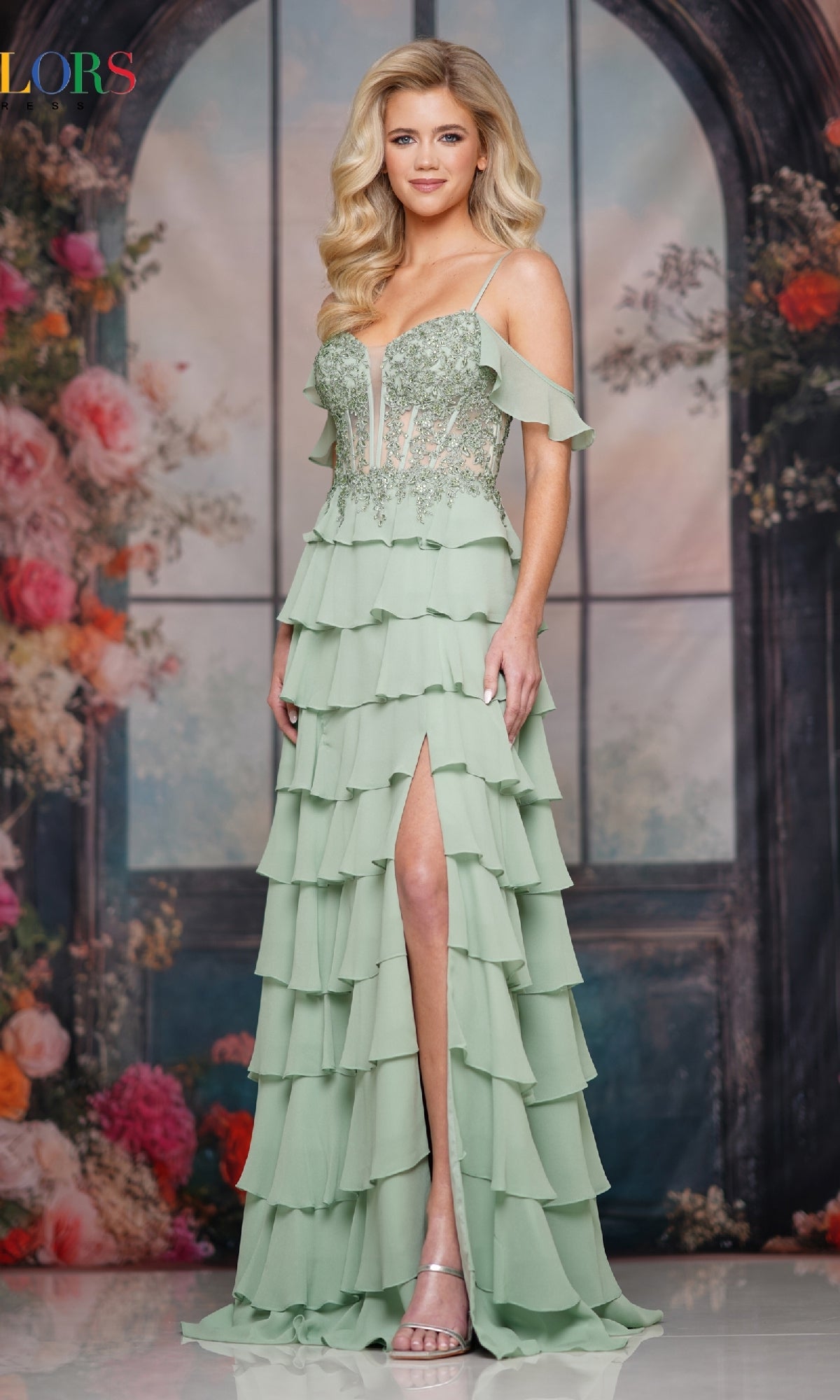 Woman wearing Colors Dress 3820, a light green long prom dress with tiered ruffles, corset bodice, off-shoulder straps, and thigh-high slit, stands before a floral backdrop.