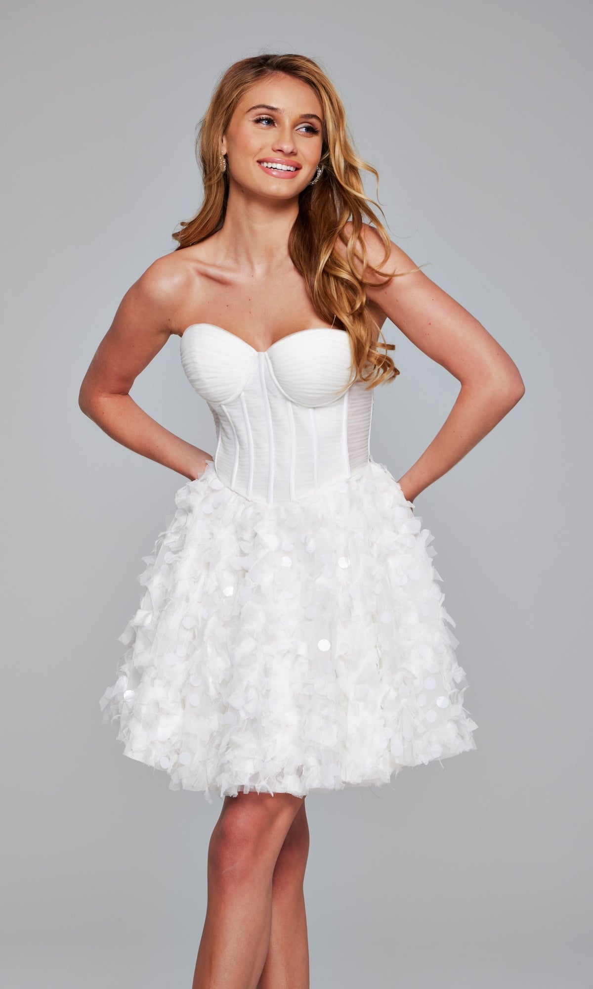 A woman wears the Jovani Strapless Short White Hoco Dress 38248, featuring a fitted bodice and a textured floral-patterned skirt, standing against a plain gray background.