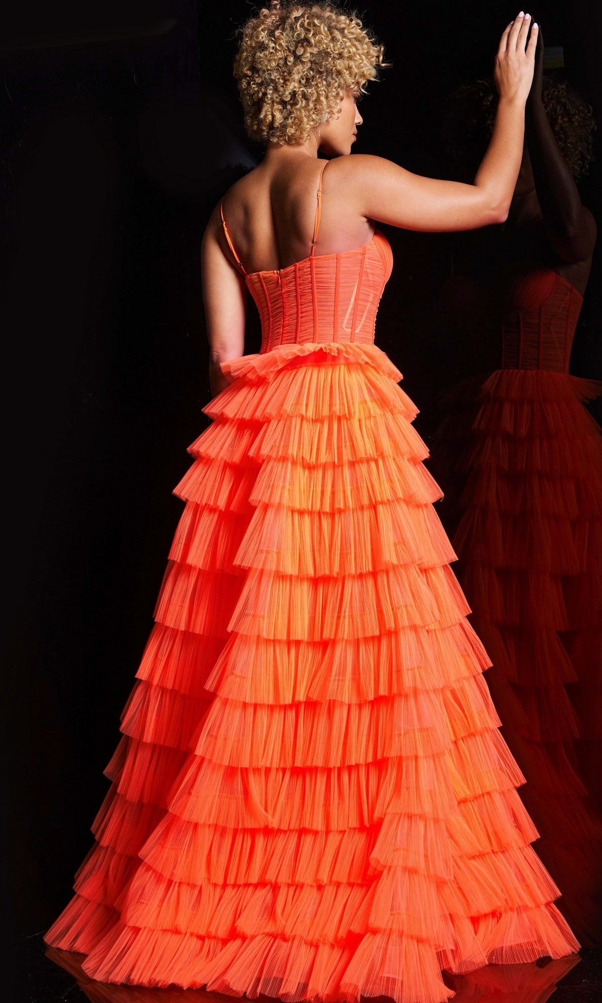 A woman with curly blonde hair, seen from behind, touches a reflective surface while wearing the Jovani Long Ruffled Corset Prom Ball Gown 38277 in bright orange.