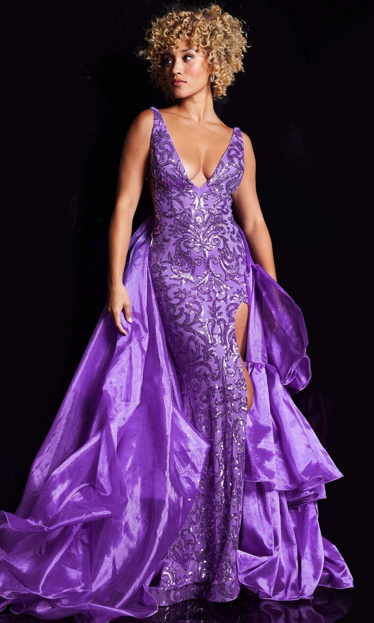 A person wearing the Jovani Ruffled Long Sequin-Print Prom Dress 38336, a fitted sleeveless gown with glittering sequin patterns and a dramatic ruffled train, stands against a black background.
