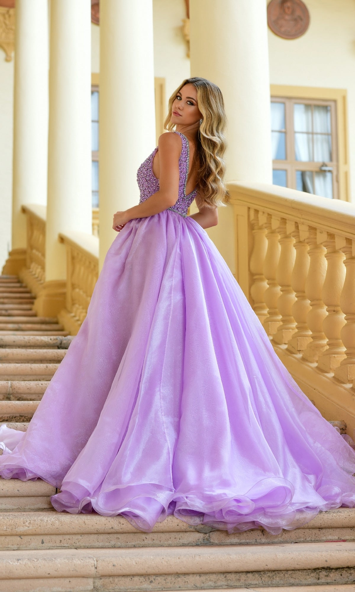 A woman in the Ava Presley 38342 Deep V-Neck Long Prom Ball Gown poses on marble stairs framed by cream columns, glancing over her shoulder. Her sleeveless lavender gown features a fitted bodice and a voluminous skirt.