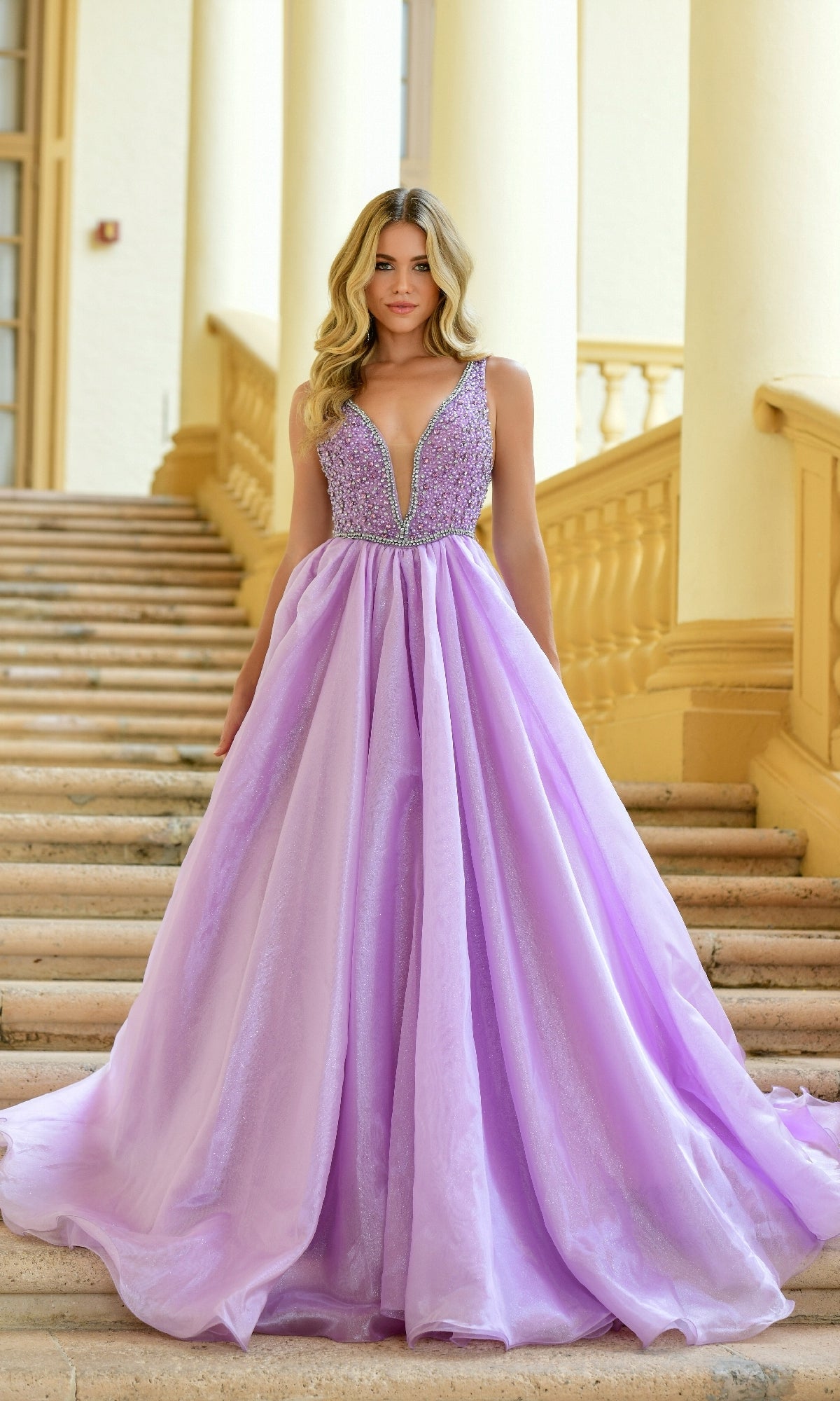 A woman stands on stone stairs wearing the Ava Presley 38342 deep V-neck long prom ball gown—a sleeveless lavender dress featuring a beaded bodice.