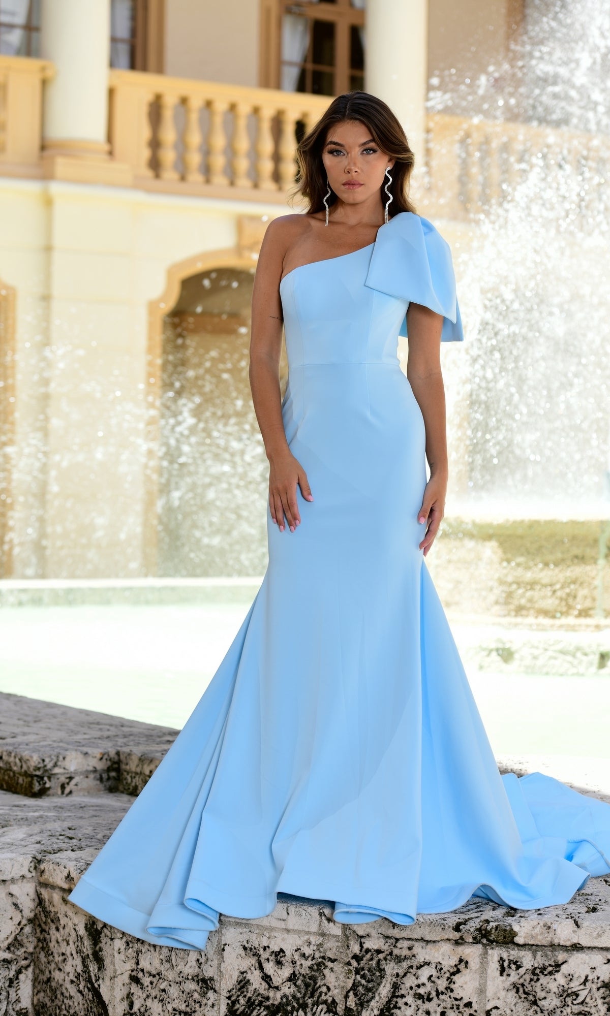 A woman in the Ava Presley 38347 One-Shoulder Mermaid Prom Gown, light blue, stands before a stone fountain and a yellow building with columns and balconies.