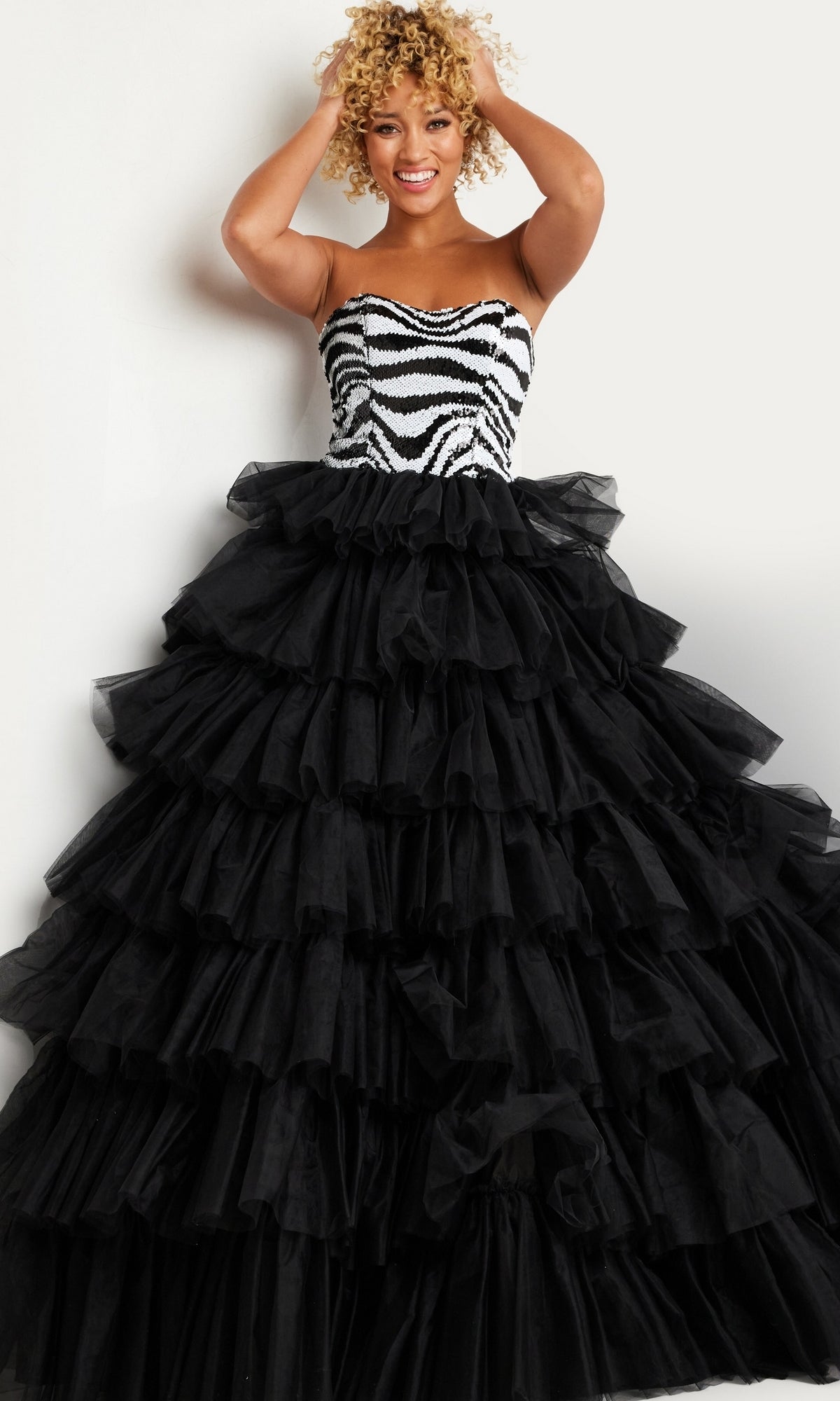A woman smiles with her hands in her hair, wearing the Jovani Black White Long Ruffled Prom Dress 38360, which features a zebra-patterned bodice and a voluminous layered tulle skirt, standing against a plain white background.