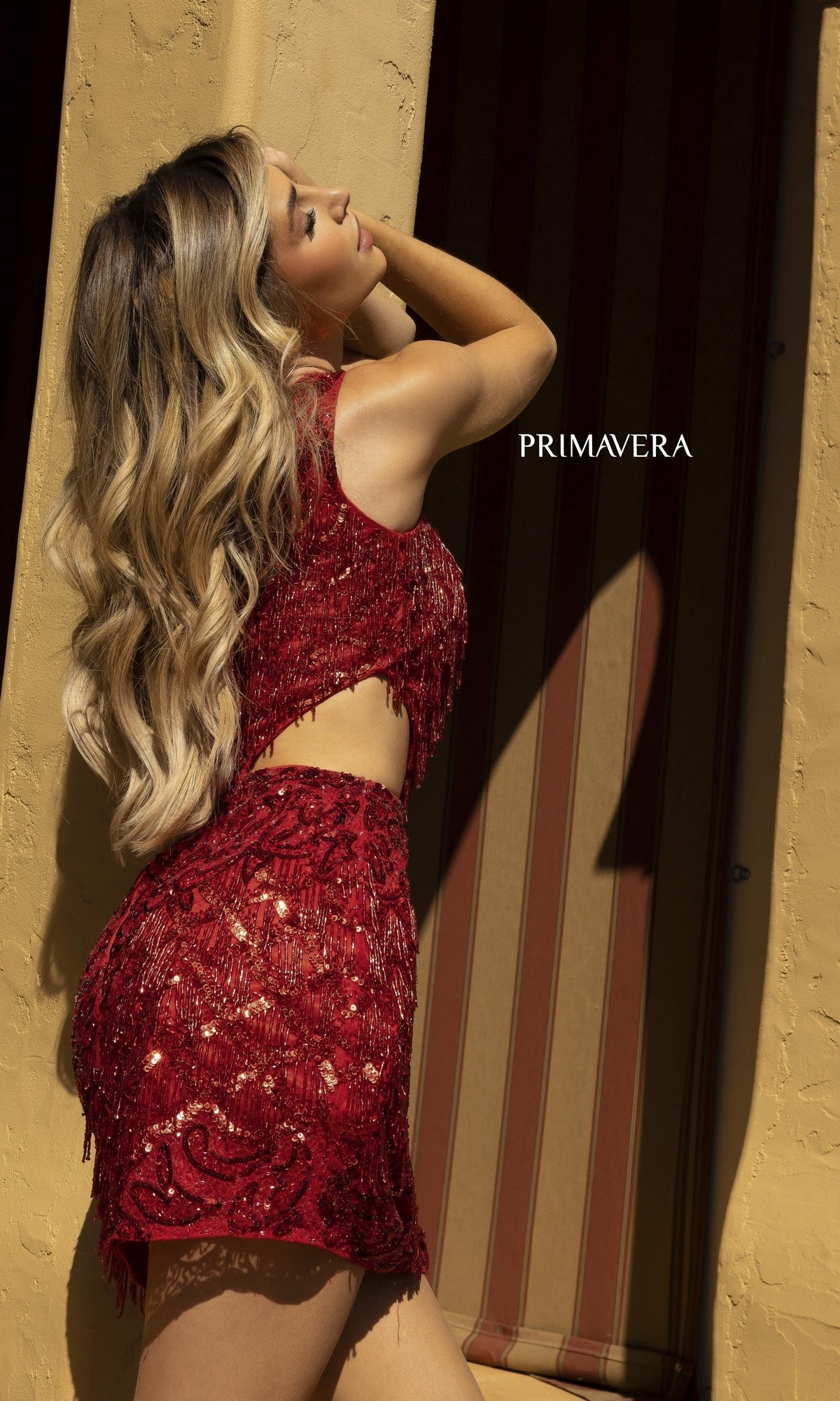 Woman with long wavy hair wearing the One-Shoulder Fringe Short Sequin Party Dress 3836, leaning against a yellow wall with