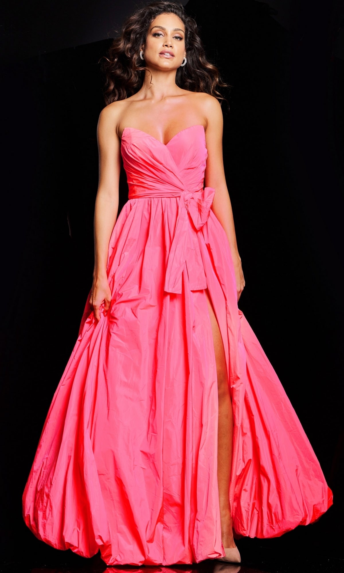 Woman in a strapless, bright pink Jovani Long Taffeta Bubble Prom Ball Gown 38382 with a floor-length skirt, bow waist detail, and high slit, posing against a black background.