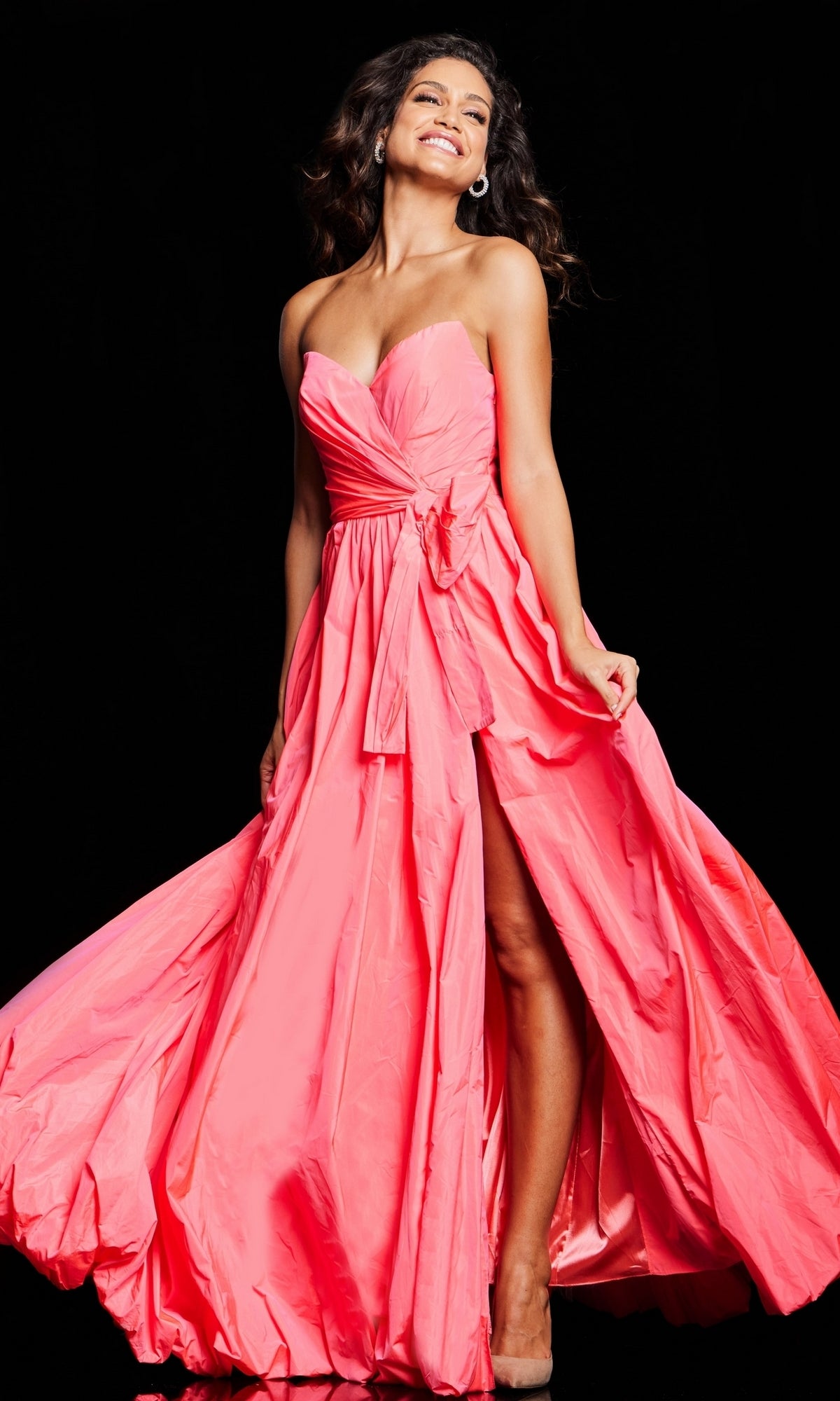 A woman in the Jovani Long Taffeta Bubble Prom Ball Gown 38382, a bright pink strapless dress with a high slit, stands smiling against a black background.