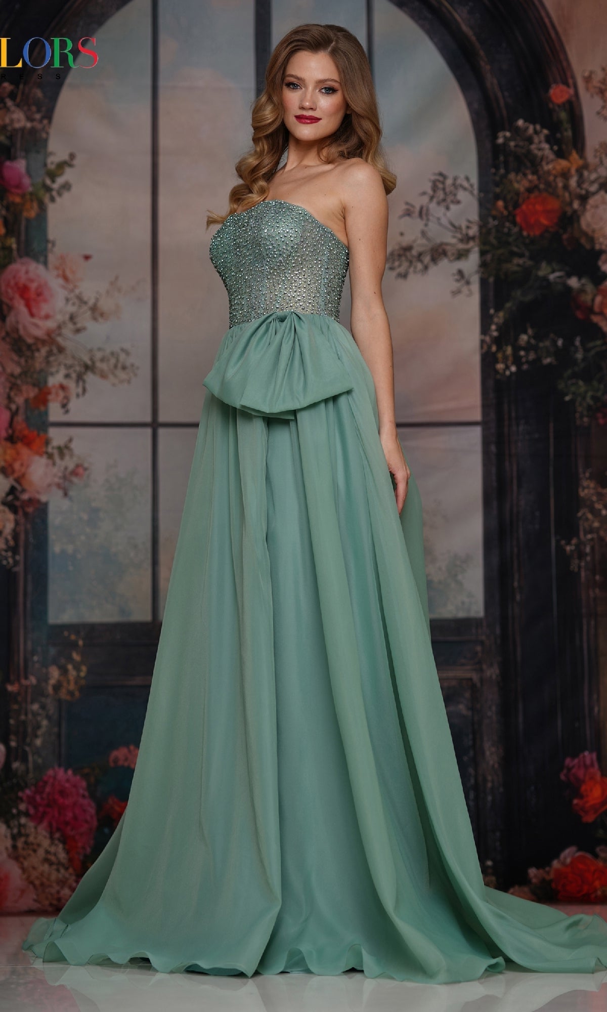 A woman stands indoors wearing the Colors Dress 3843, a strapless long prom dress with a hot-stone embellished bodice and flowing chiffon A-line skirt; floral and window backdrop.