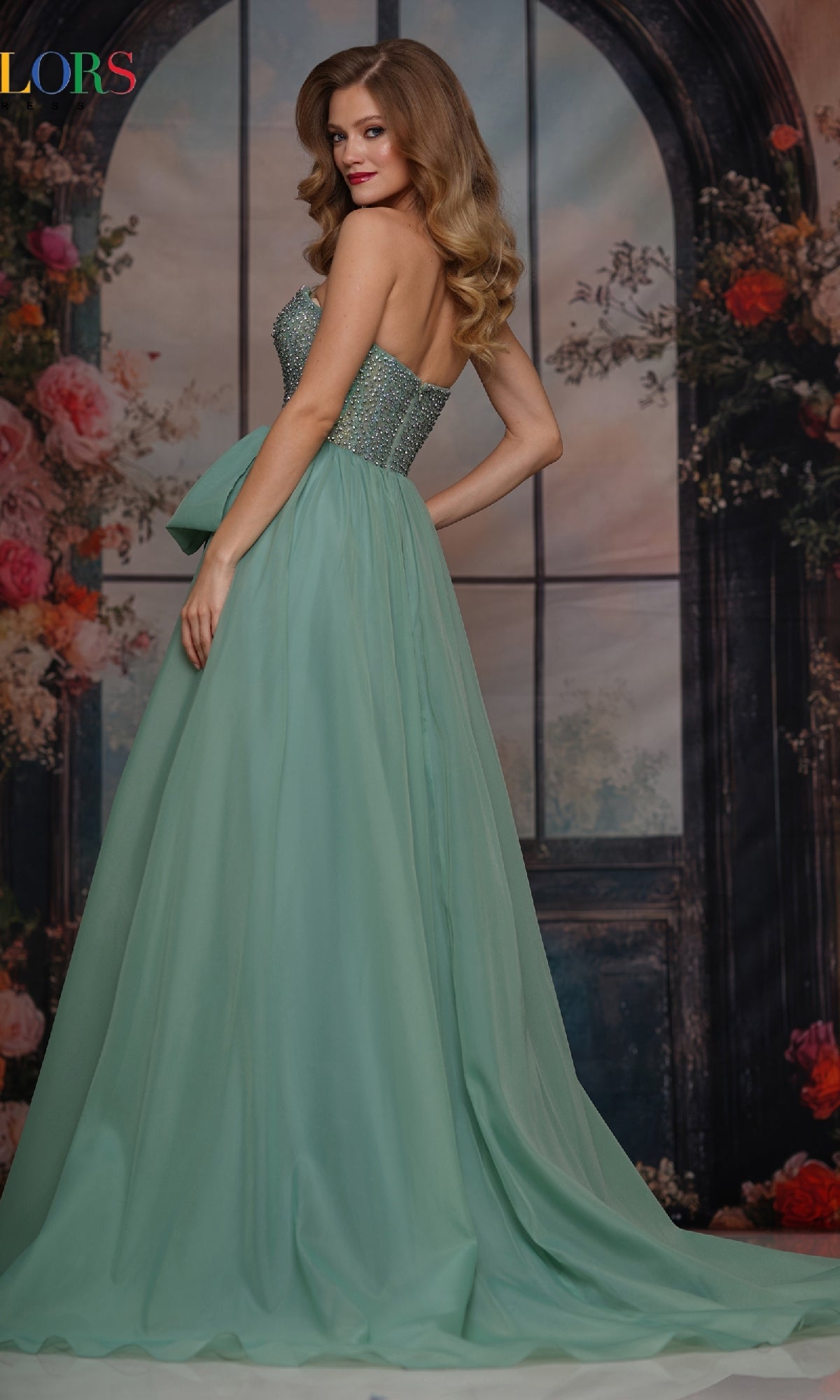 A woman poses before a floral backdrop, looking over her shoulder in the Colors Dress 3843, a long prom dress with a strapless hot-stone embellished bodice and flowing chiffon A-line skirt.