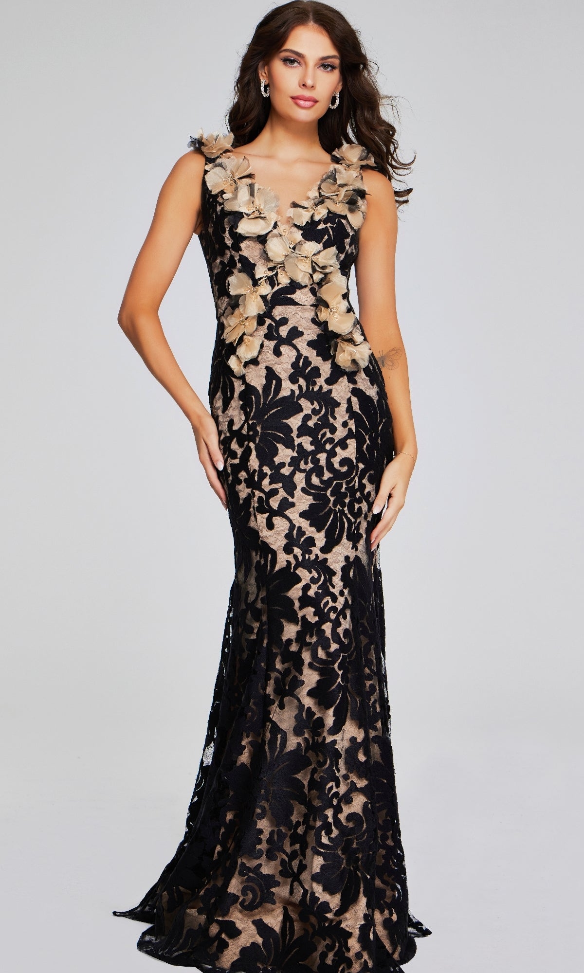 A woman stands against a light background wearing the Jovani 38497 Long Black & Nude Formal Dress, a sleeveless, floor-length gown featuring a beige base, black lace overlay, and 3D flower accents on the neckline.