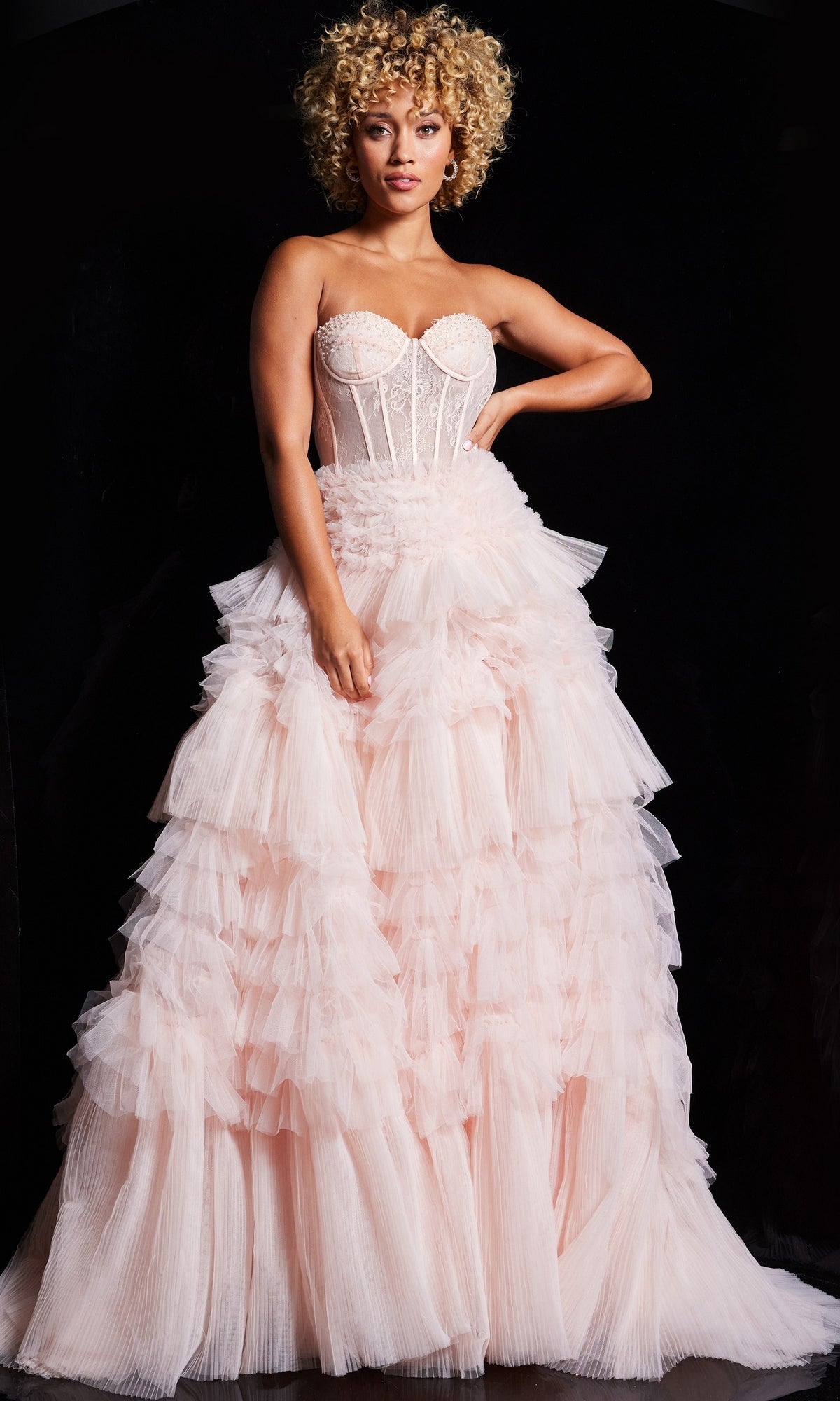 A woman with curly blonde hair wears the Jovani Ruffled Long Strapless Prom Ball Gown 38540 in light pink, featuring a fitted bodice and tiered pleated skirt, standing against a dark background.