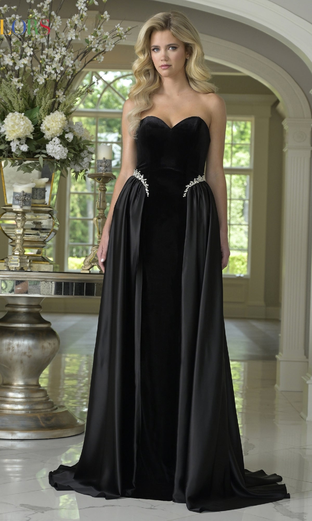 A woman wears the Strapless Velvet Prom Gown: Colors Dress 3861, featuring crystal-beaded accents, while standing indoors in a bright, elegantly decorated room with flowers.