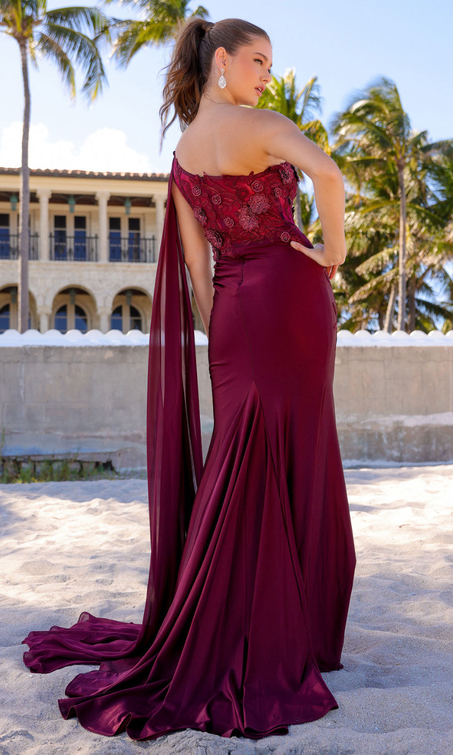 Woman standing on sand in front of a building framed by palm trees, wearing a burgundy One-Shoulder Long Formal Dress with Cape 388, featuring a corset-style bodice and flowing train under a clear blue sky.