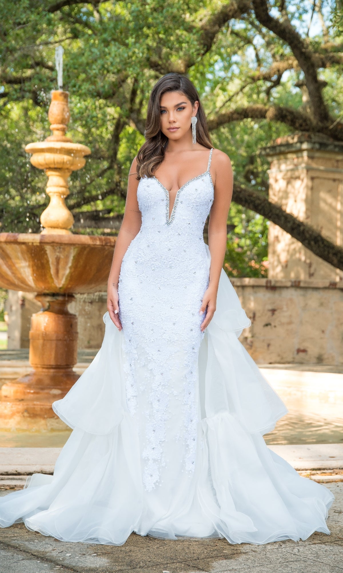 A woman stands outdoors in the Long Formal Dress 38813 by Ava Presley—a fitted white gown with a deep V-neckline, embroidered corset bodice, beaded accents, and sheer train—near a fountain and trees.