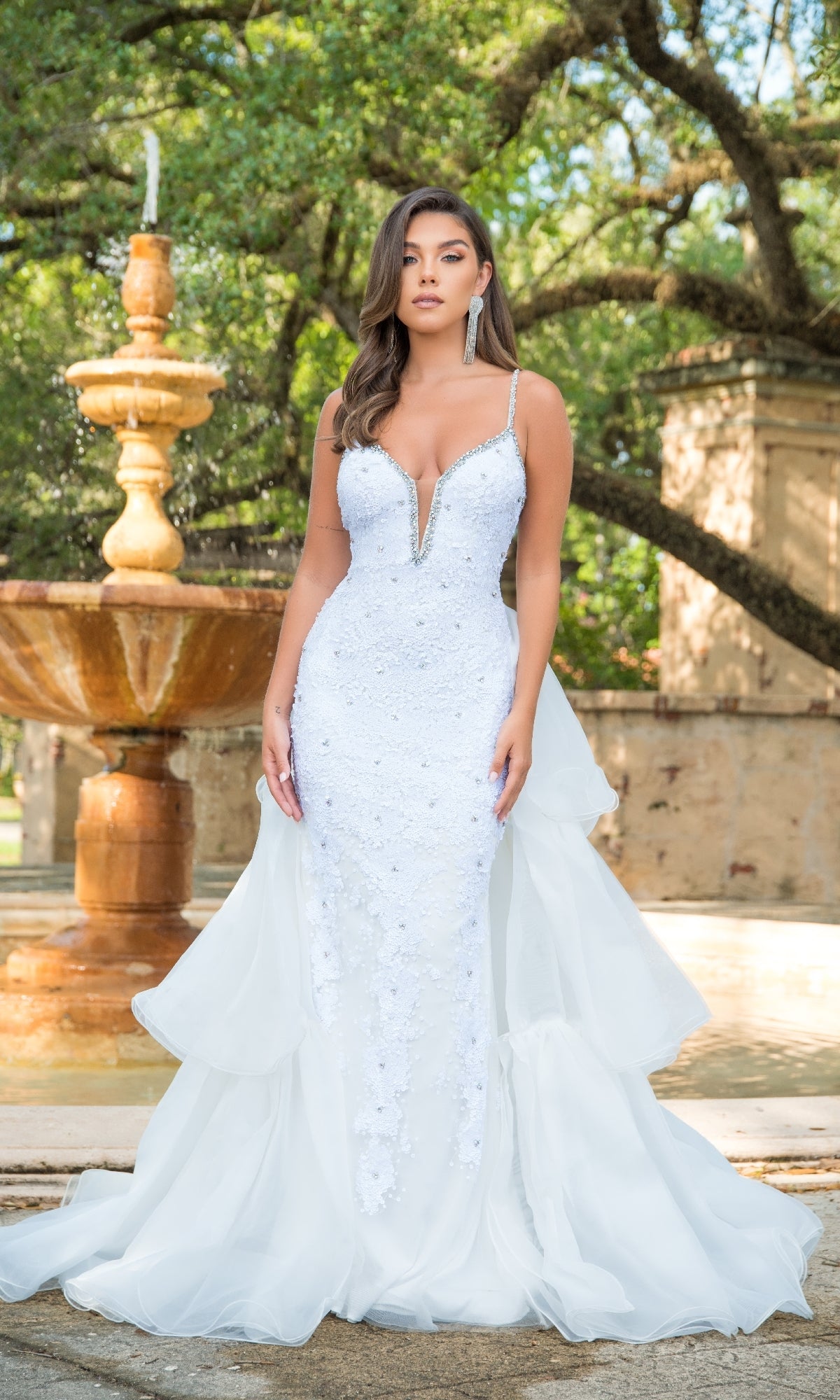 A woman stands before a fountain and trees wearing the Long Formal Dress 38813 by Ava Presley, featuring a fitted white silhouette with an embroidered corset bodice and deep neckline.