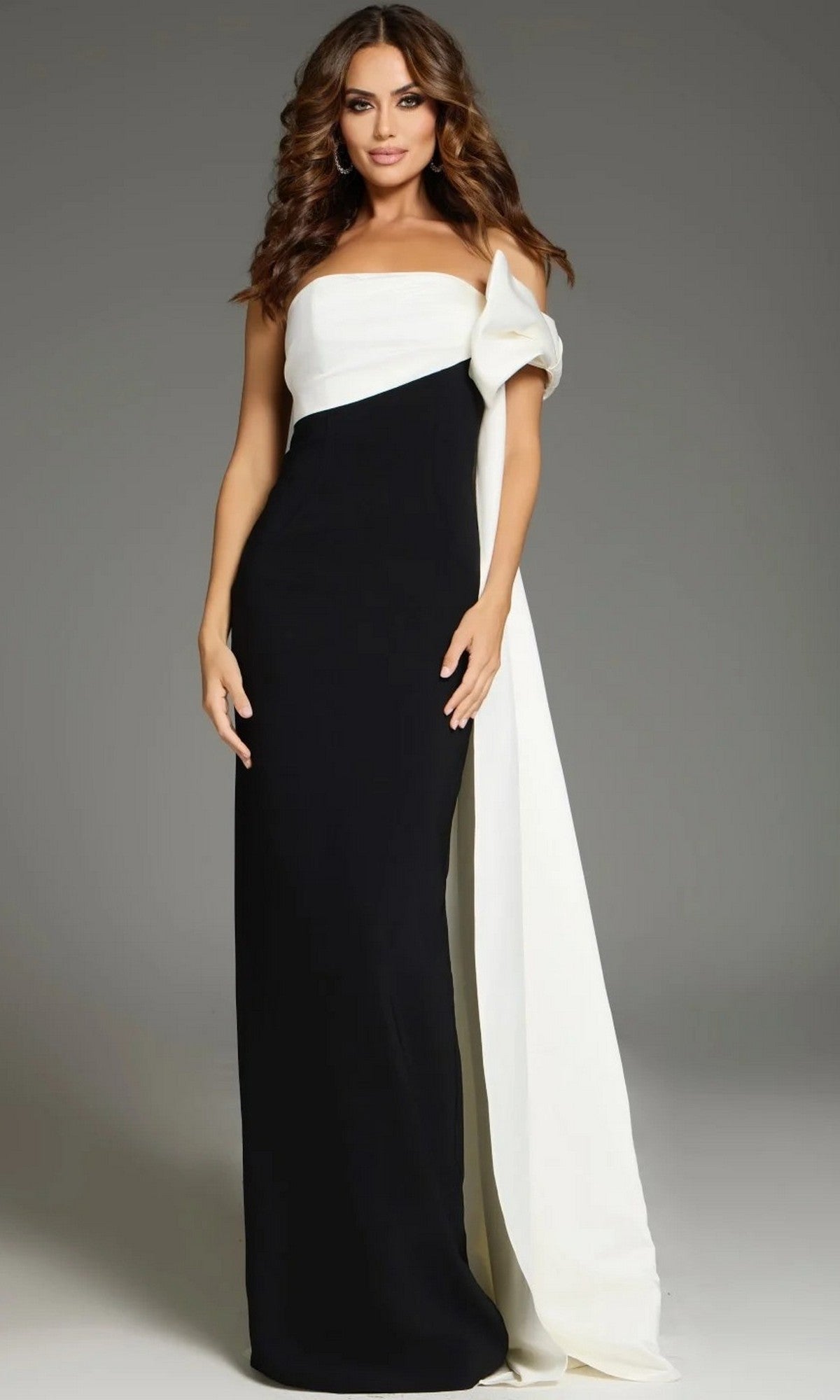 A woman stands against a plain gray background wearing the Jovani 38864 Long Formal Dress—a floor-length black gown featuring a white asymmetrical overlay and draped fabric detail over one shoulder.
