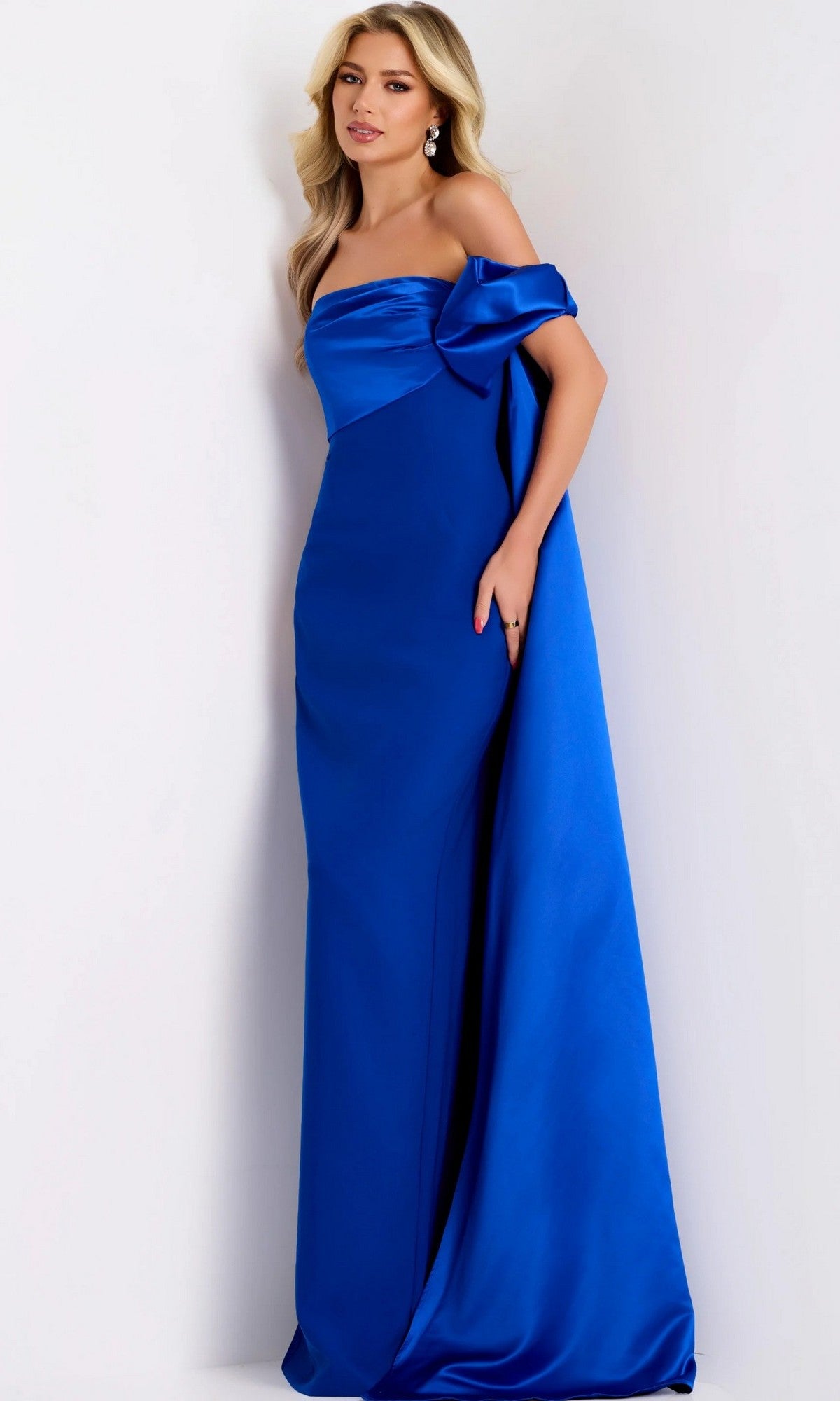 A woman stands against a white background, wearing the Jovani 38864 long formal dress in royal blue—an off-the-shoulder gown featuring draped detailing—paired with matching earrings.