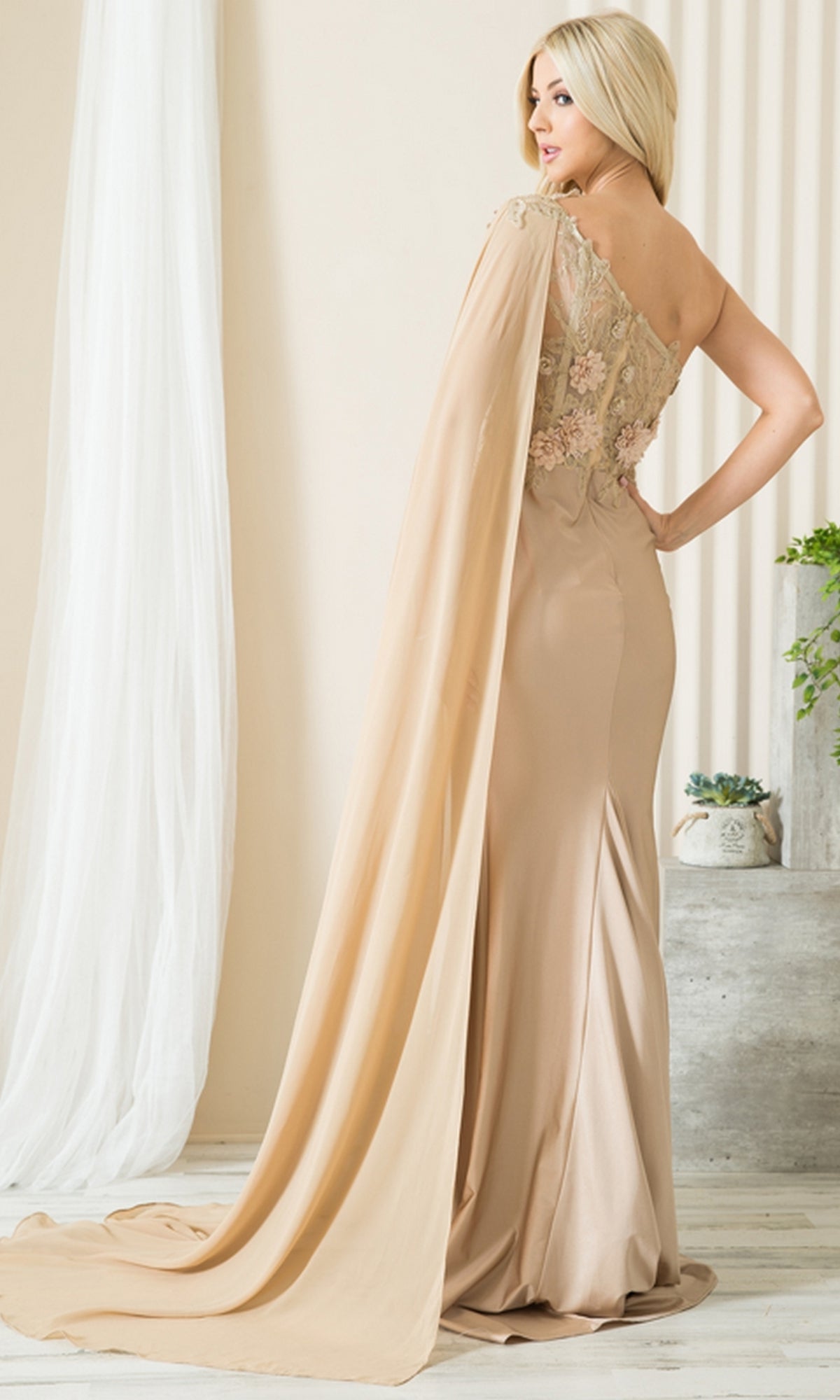 A woman stands indoors wearing the One-Shoulder Long Formal Dress with Cape 388, featuring a corset-style bodice, floral embroidery, and a draped cape, looking elegantly over her shoulder.