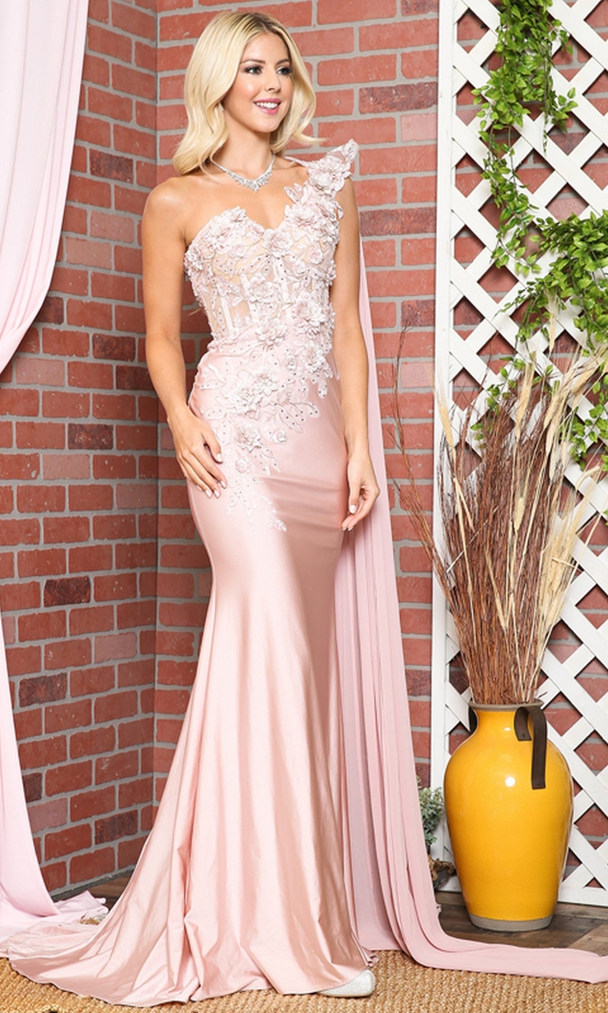 Woman wearing a light pink One-Shoulder Long Formal Dress with Cape 388, featuring a corset-style bodice and floral lace details, stands beside a yellow vase in front of a brick wall and lattice backdrop.