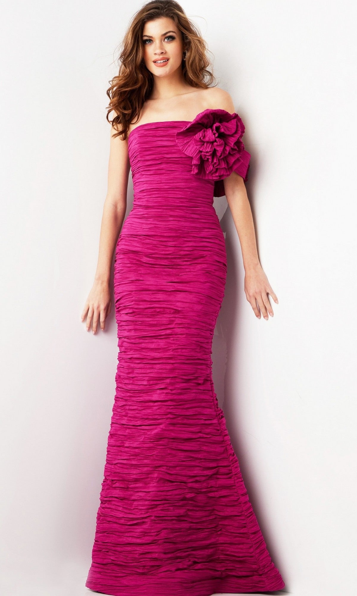 A woman poses against a plain white background wearing the Jovani 38947 long formal dress in fuchsia, featuring a fitted, strapless design with ruched texture and a prominent fabric flower detail on the left shoulder.