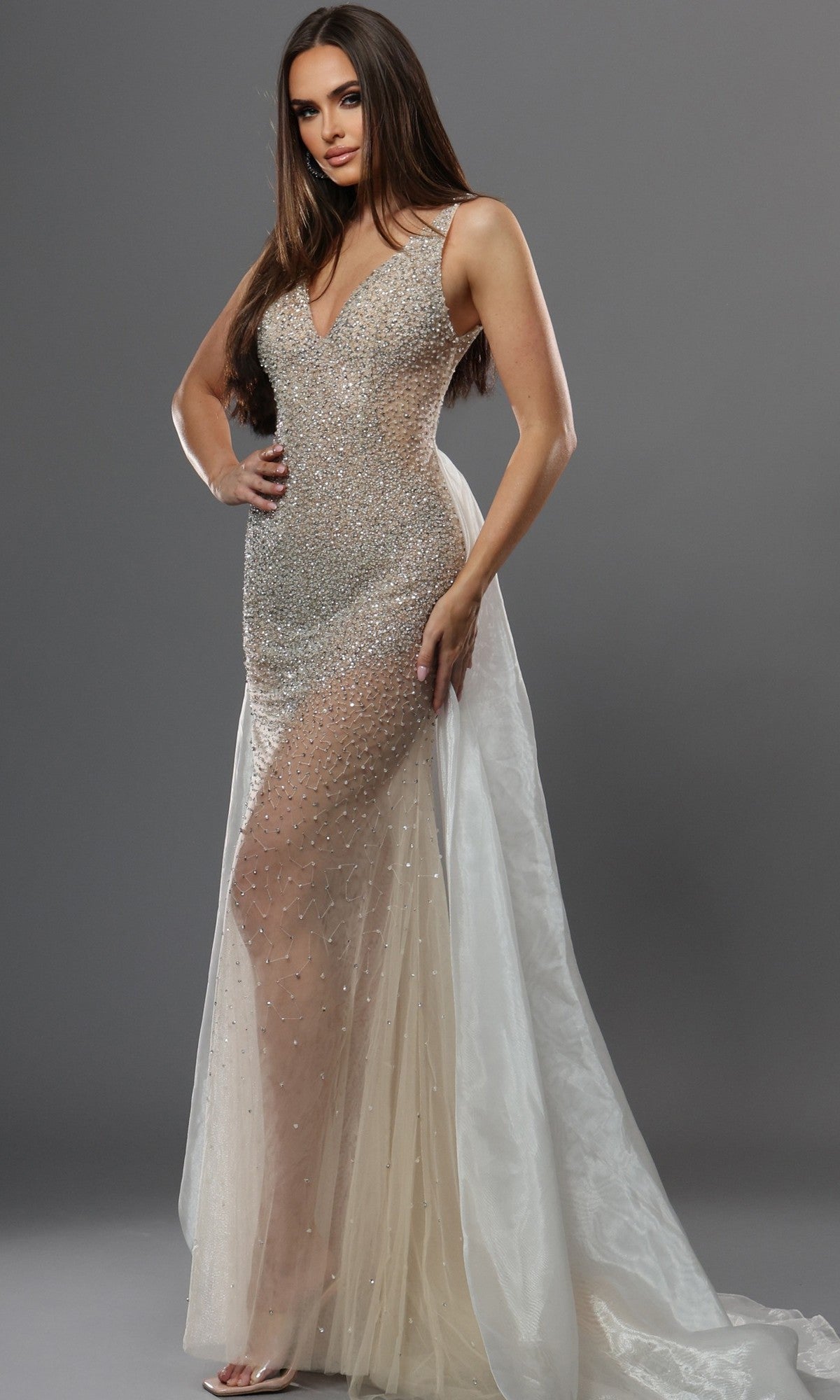 A woman wearing the Jovani 39043 long formal dress in nude/off white—a sleeveless, floor-length gown adorned with crystal embellishments, featuring sheer panels and a long, flowing train—poses against a plain gray background.