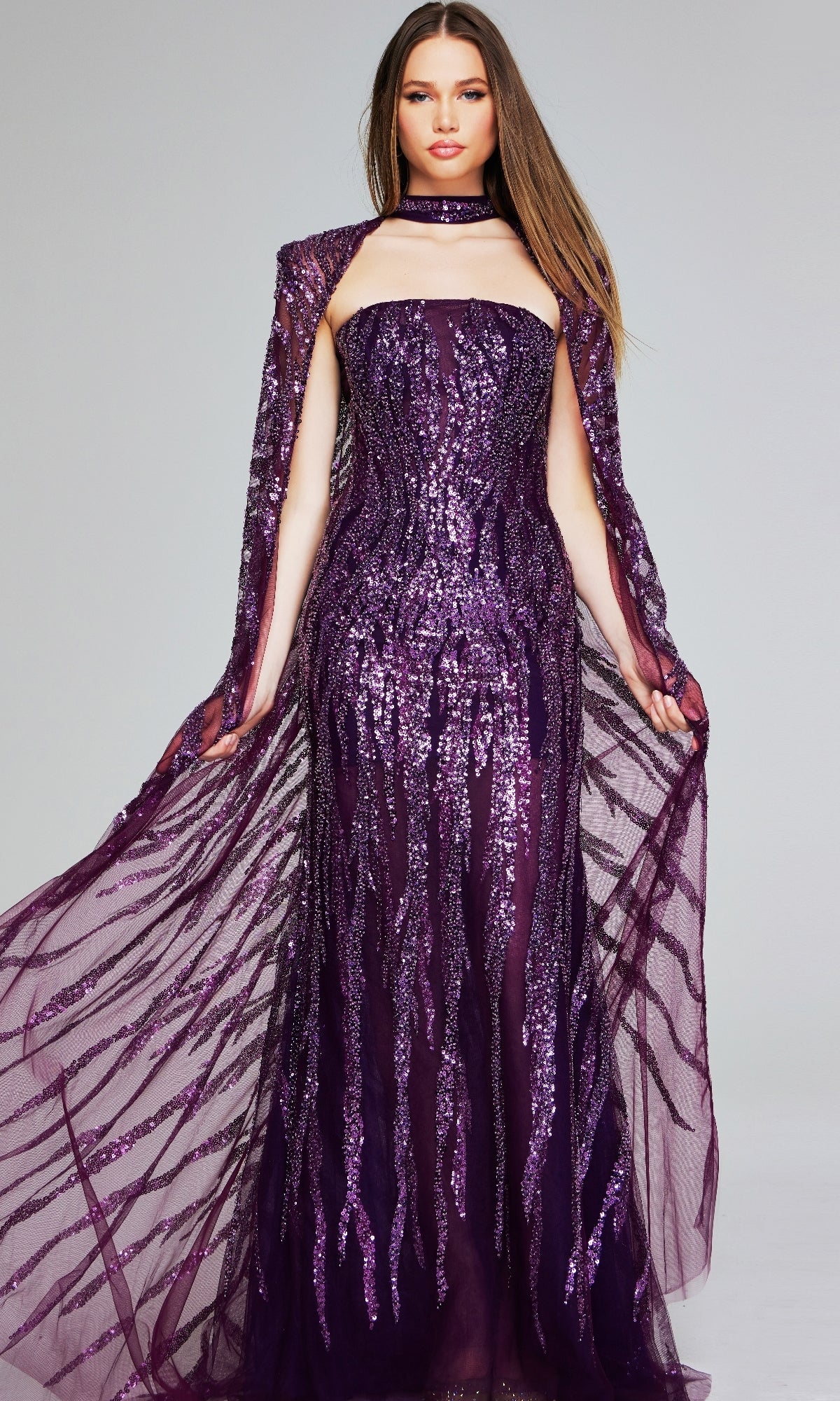 A woman models the Jovani 39046 Purple Sequin Pageant Dress—floor-length, strapless, with a matching sheer cape and choker—against a plain background, making it an elegant pick for any special occasion.